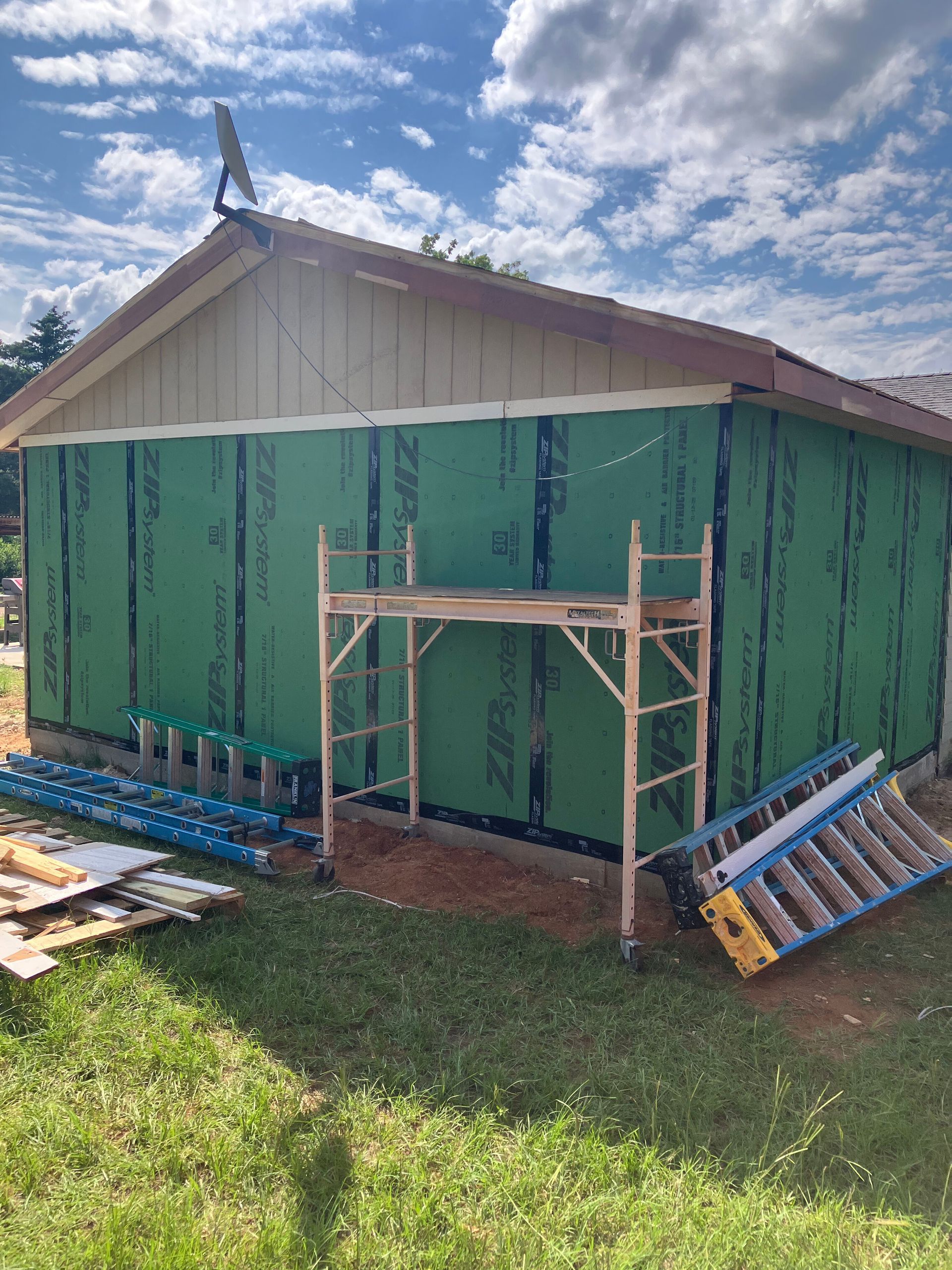 A building exterior covered in green structural sheathing with scaffolding and ladders set up in front on a sunny day.