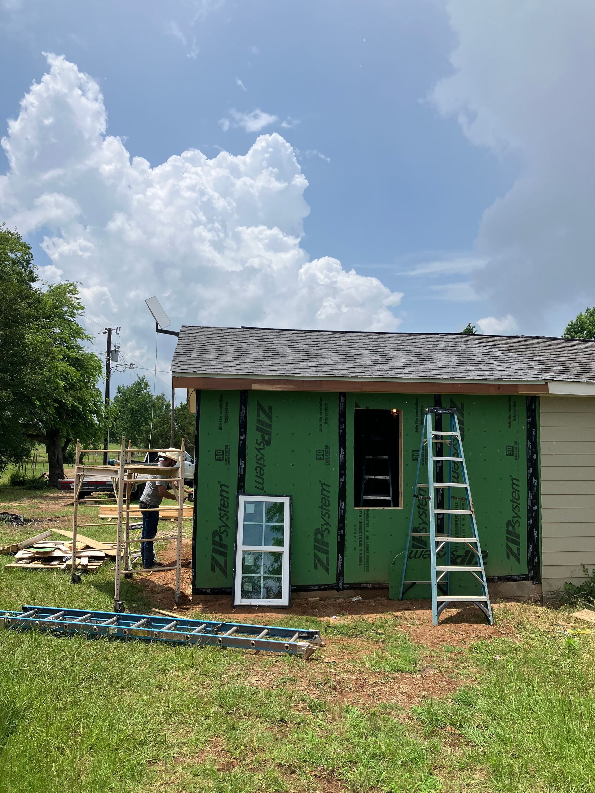 A person works on a shed frame covered in green Zip System panels, with a window, two ladders, and a cloudy blue sky.