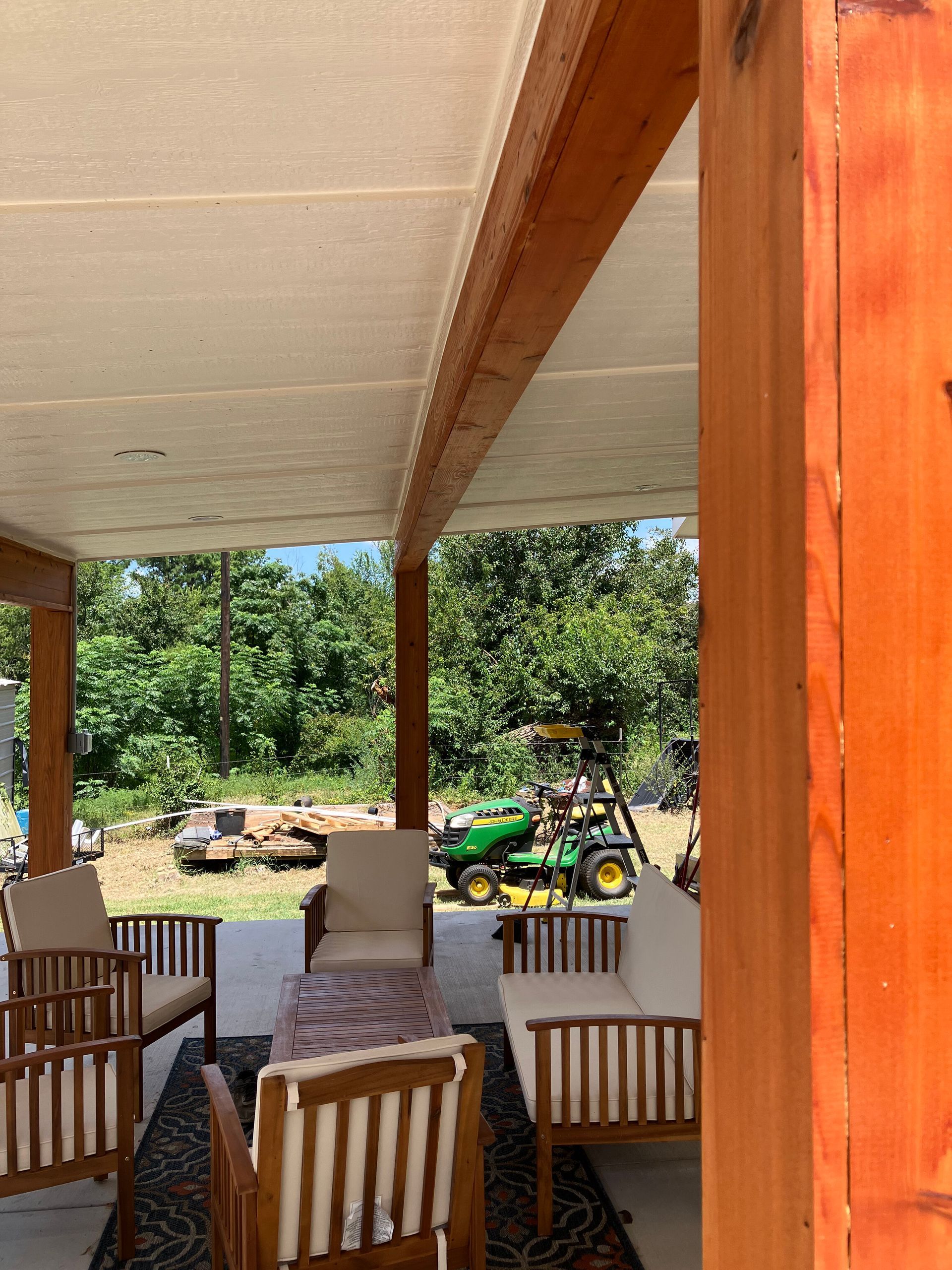 A patio with wooden chairs, a sofa, and a table underneath a covered roof, looking out onto a grassy yard with a tractor.