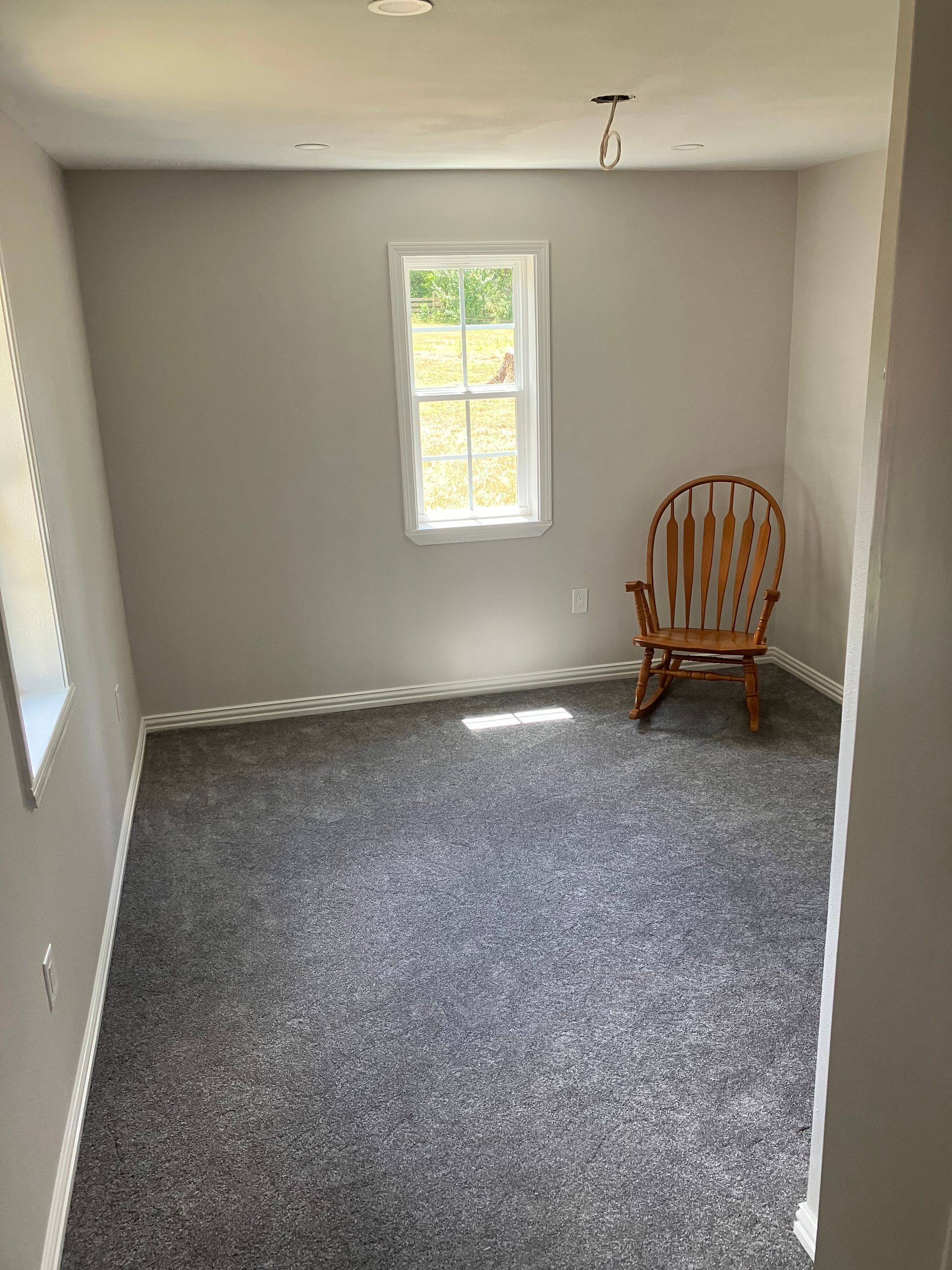 A small, empty room with grey walls, dark grey carpet, a single window, and a wooden rocking chair in the corner.