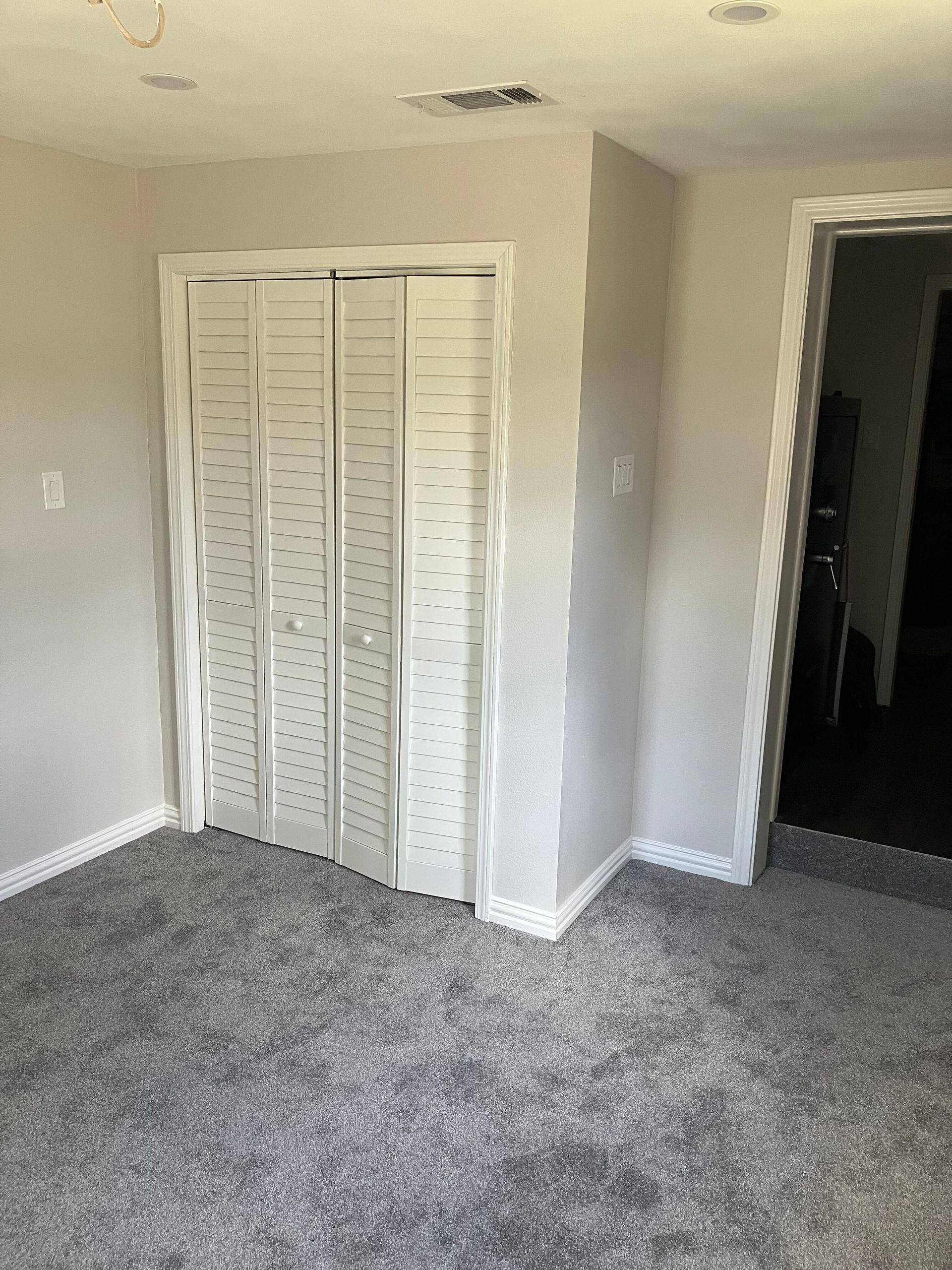 A room with beige walls and gray carpet, featuring a white louvered bifold closet door and an open doorway to another room.