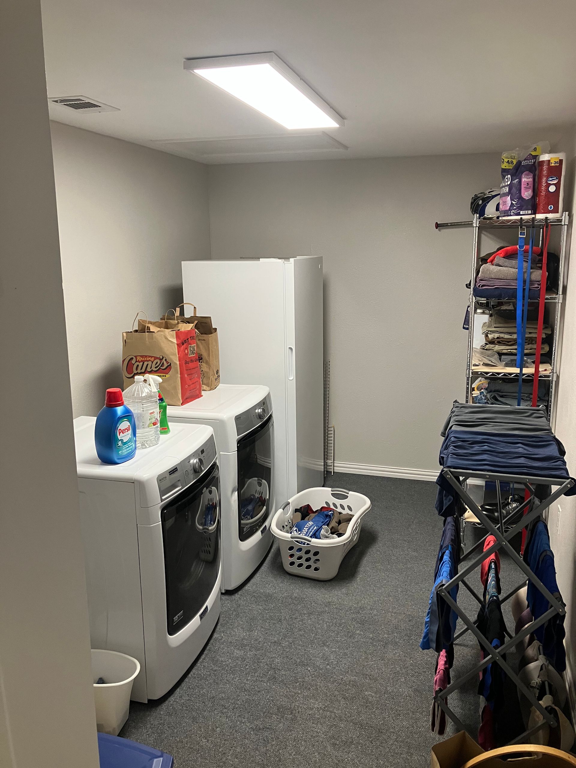 Laundry room with white washer, dryer, upright freezer, and a metal drying rack in a carpeted room.