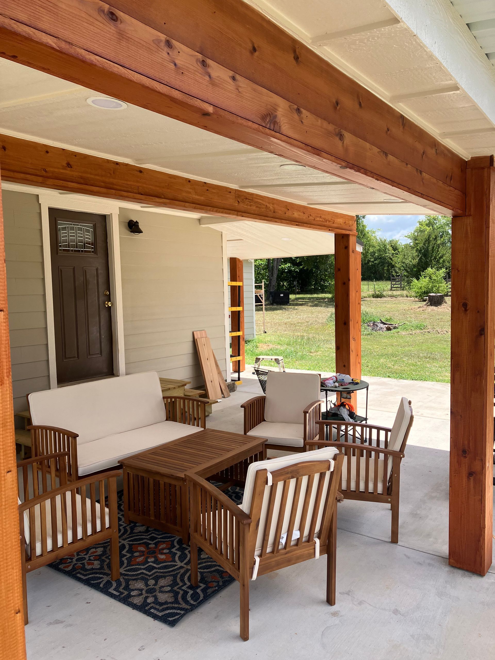 A patio with a brown wooden frame, beige sofa, chairs, and a coffee table on a patterned rug in a backyard setting.