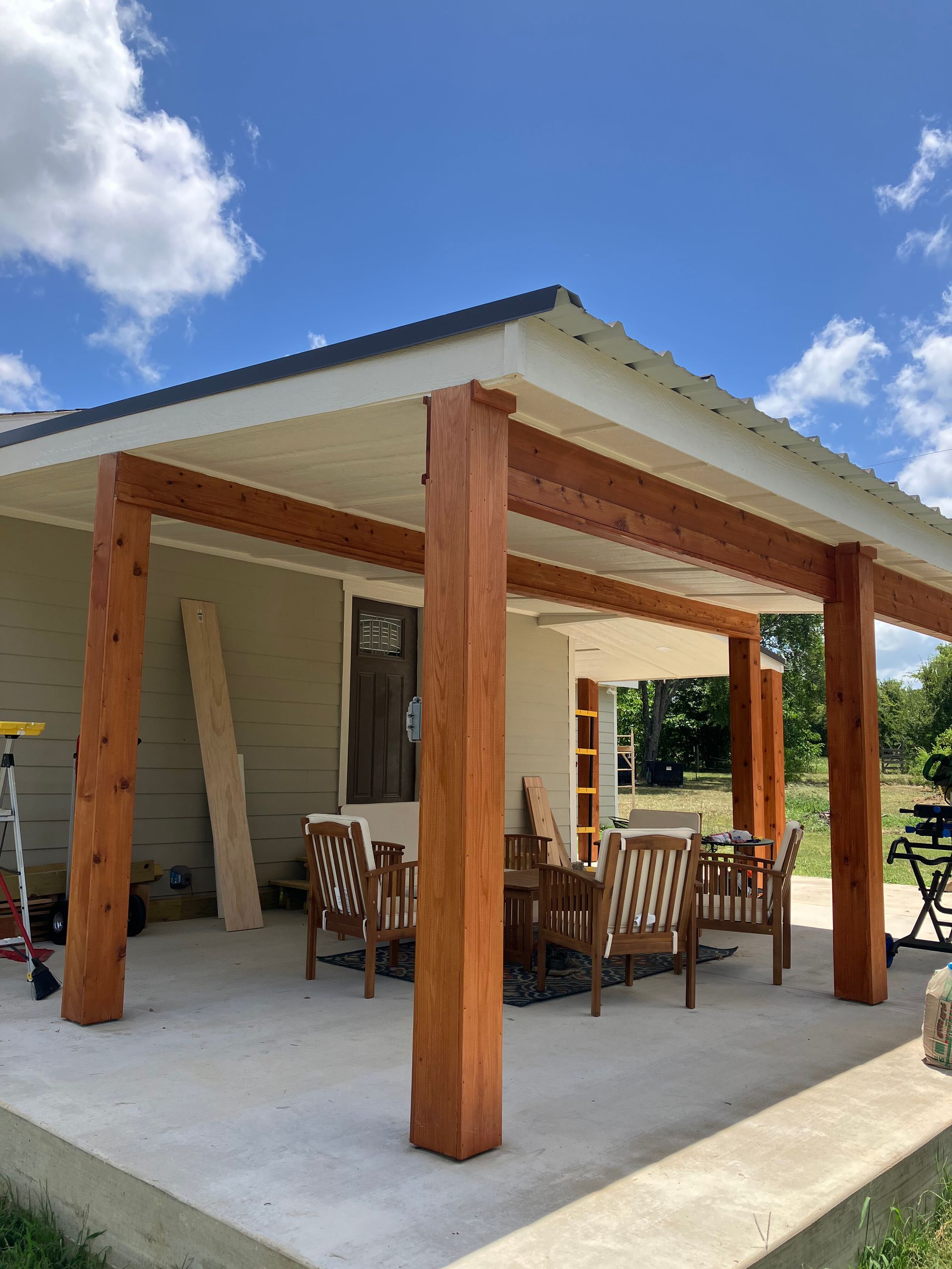 A covered concrete patio with natural wood support posts, outdoor furniture, and a house exterior under a blue sky.