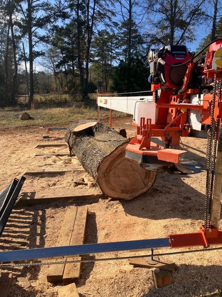 A large log sits on a portable sawmill track in an outdoor setting surrounded by trees.