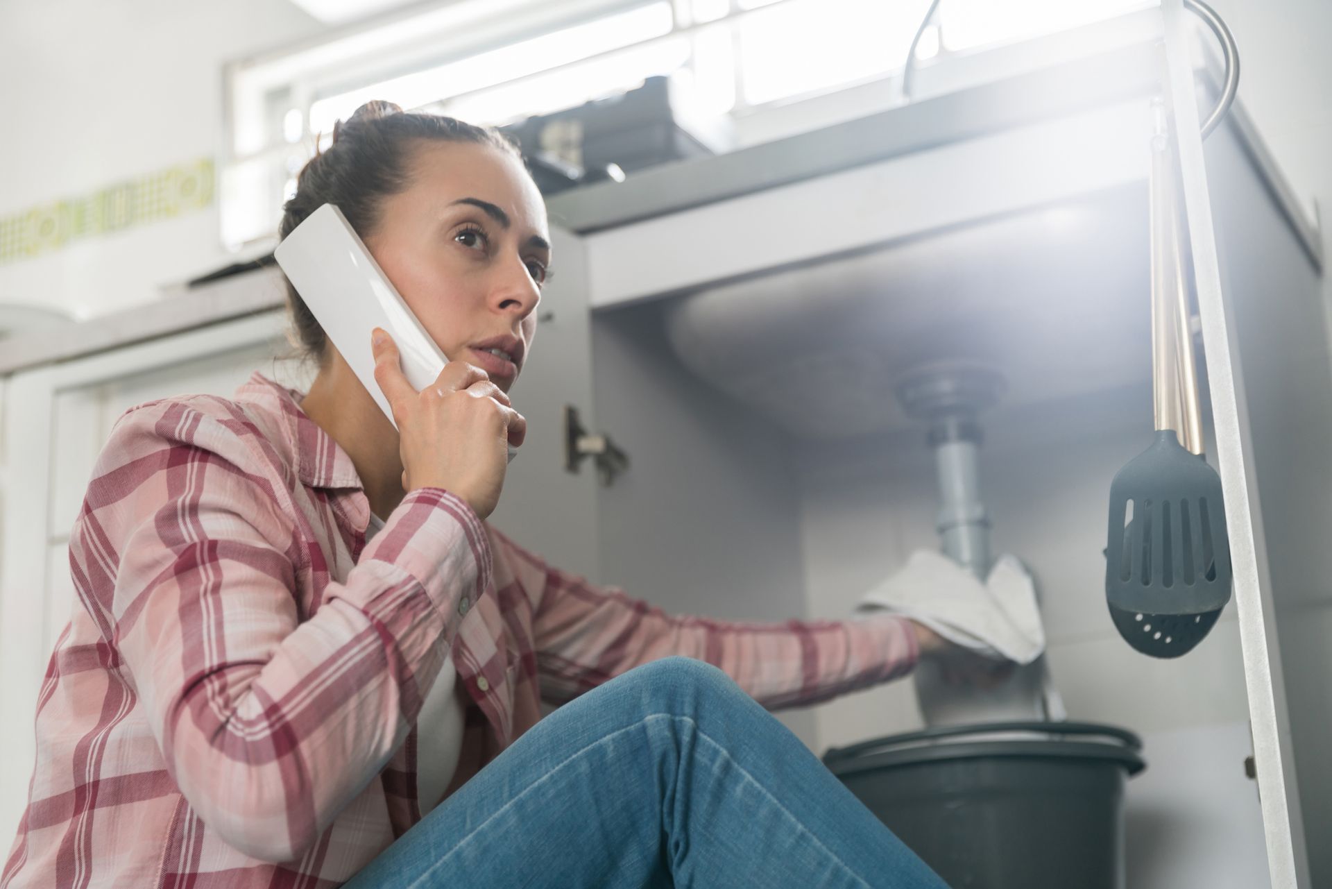 A woman is sitting under a sink talking on a cell phone.