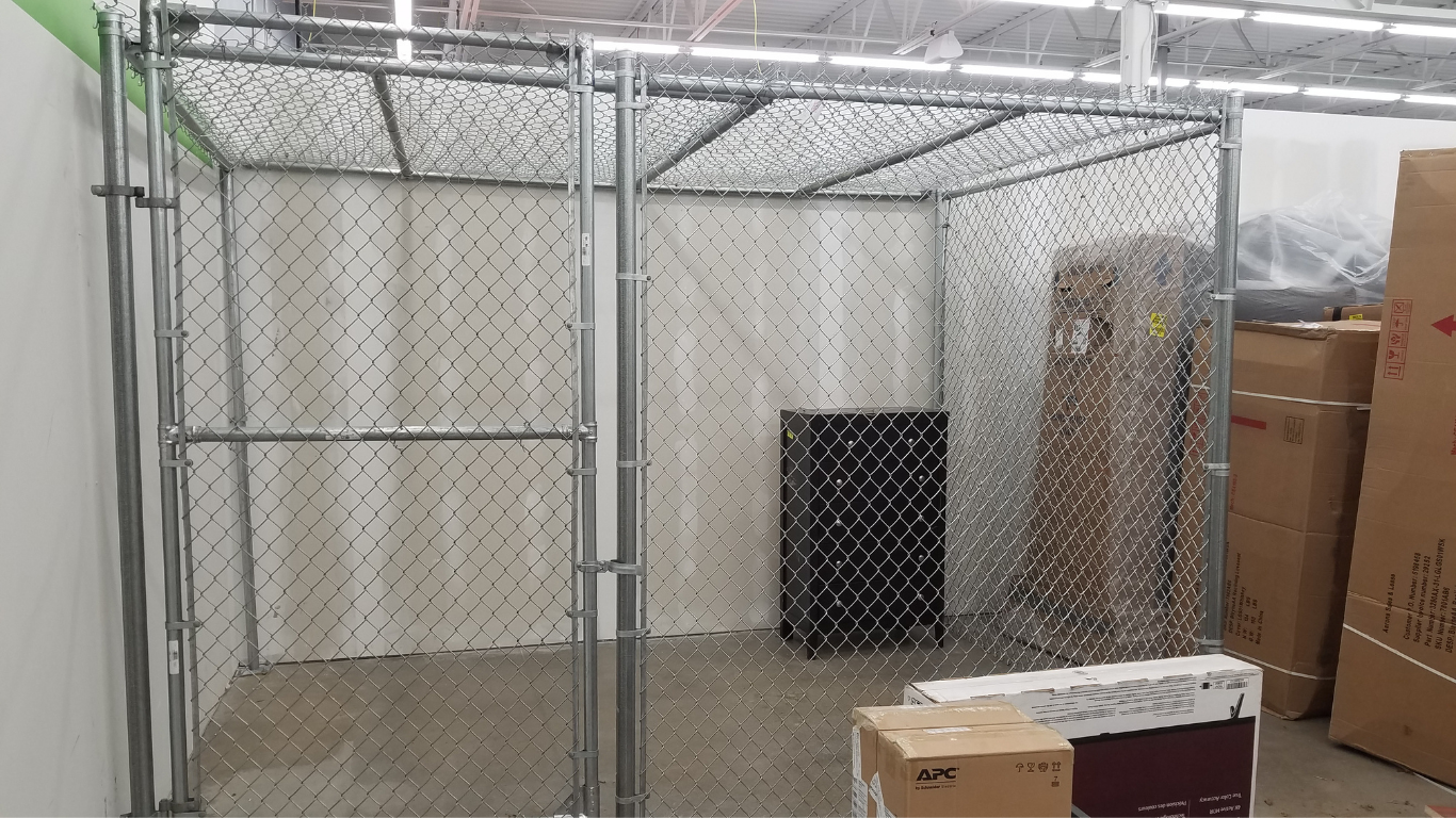 Chain-link fenced storage cage inside a store, containing a black bin and cardboard boxes.