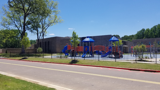 Playground next to a low brick building and a road. Blue and red play equipment with a chain link fence.