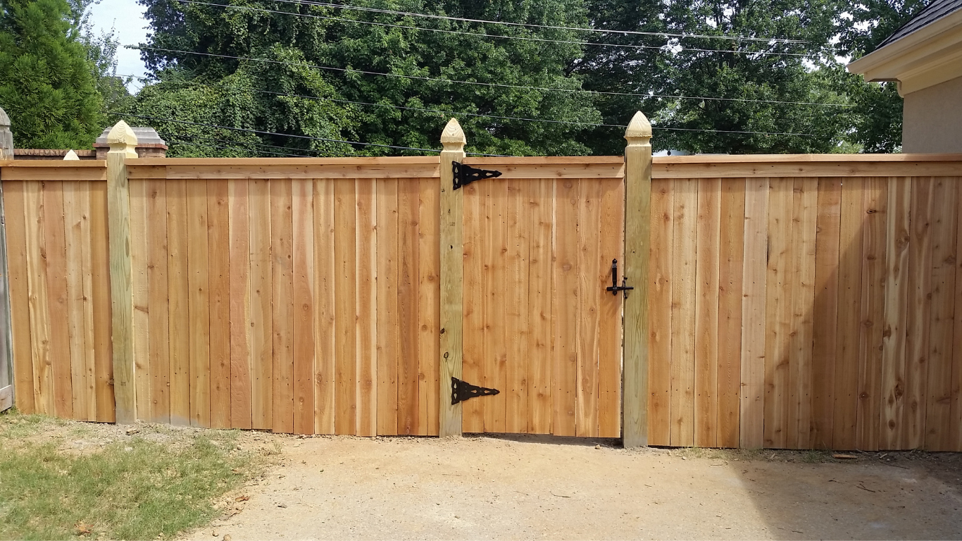 Wooden fence with gate, brown, surrounded by trees and a gravel driveway.
