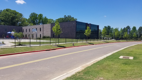 School building behind a black fence, with a road and grass in the foreground under a blue sky.