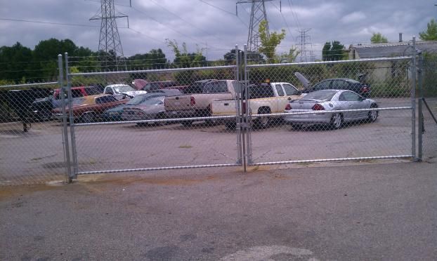 Fenced lot with several vehicles, including trucks and cars, under an overcast sky.