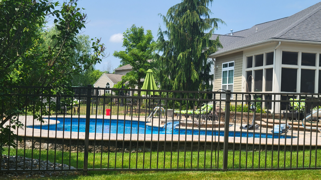 Black fenced-in backyard pool with a house in the background. Green trees and lawn under a bright blue sky.