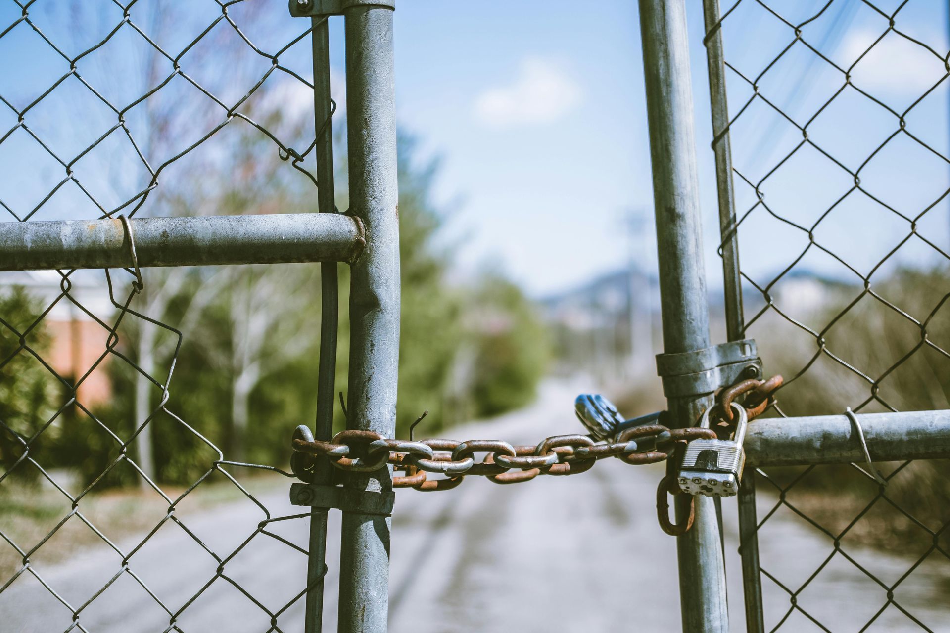 Locked chain-link gate blocks a path. Blue sky, blurred trees in background.