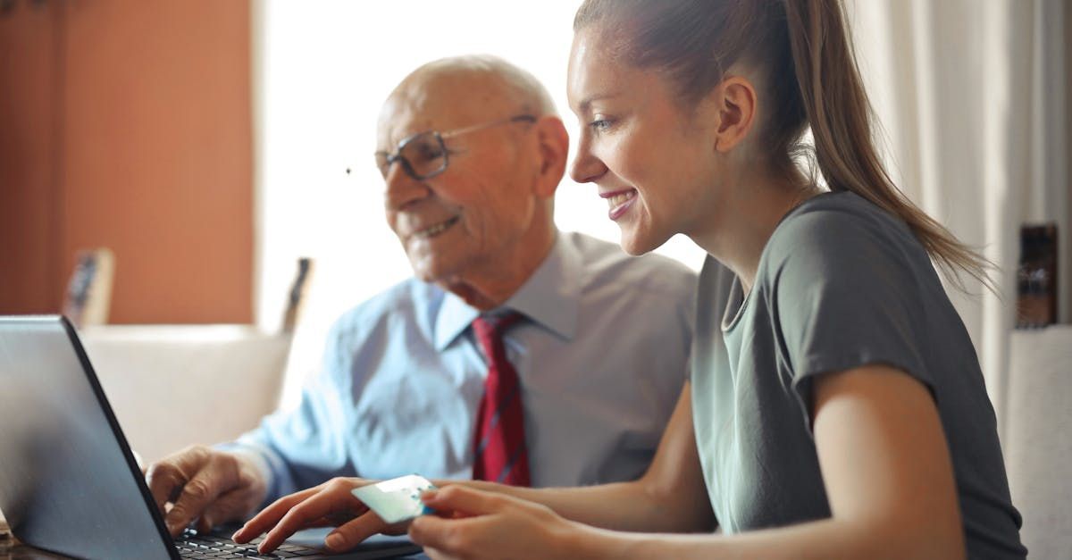 An elderly man and a young woman are sitting at a table using a laptop computer.