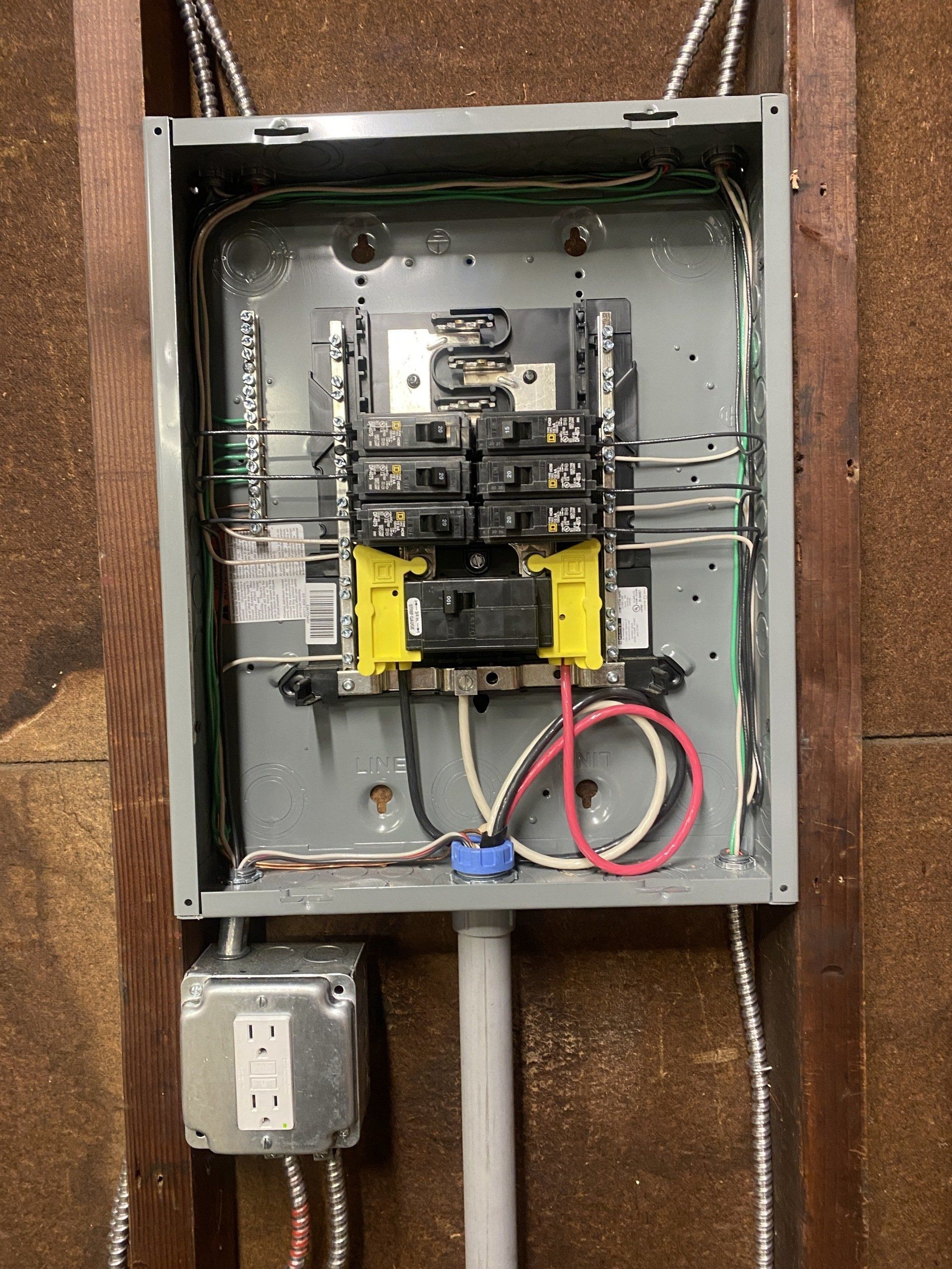 Electrical panel with circuit breakers, wires, and an outlet, mounted on a wooden wall.