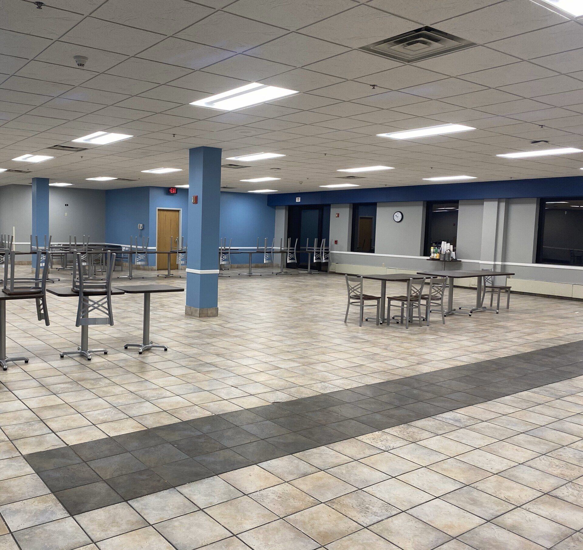 Empty cafeteria: tables, chairs, blue columns, tiled floor, fluorescent lights, pale blue walls.