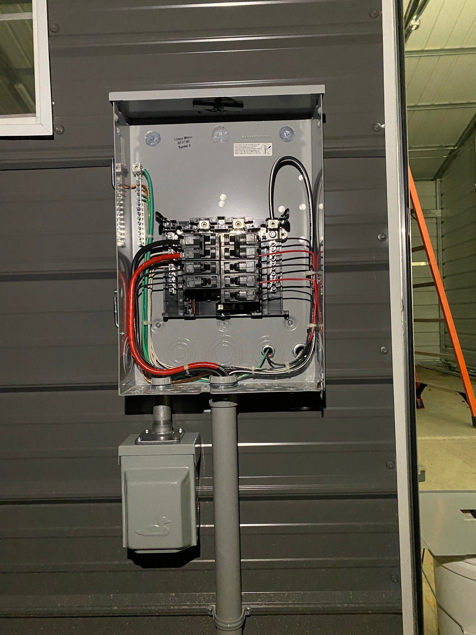Electrical panel mounted on a gray metal wall. Wires and breakers visible inside. Conduit and meter below.