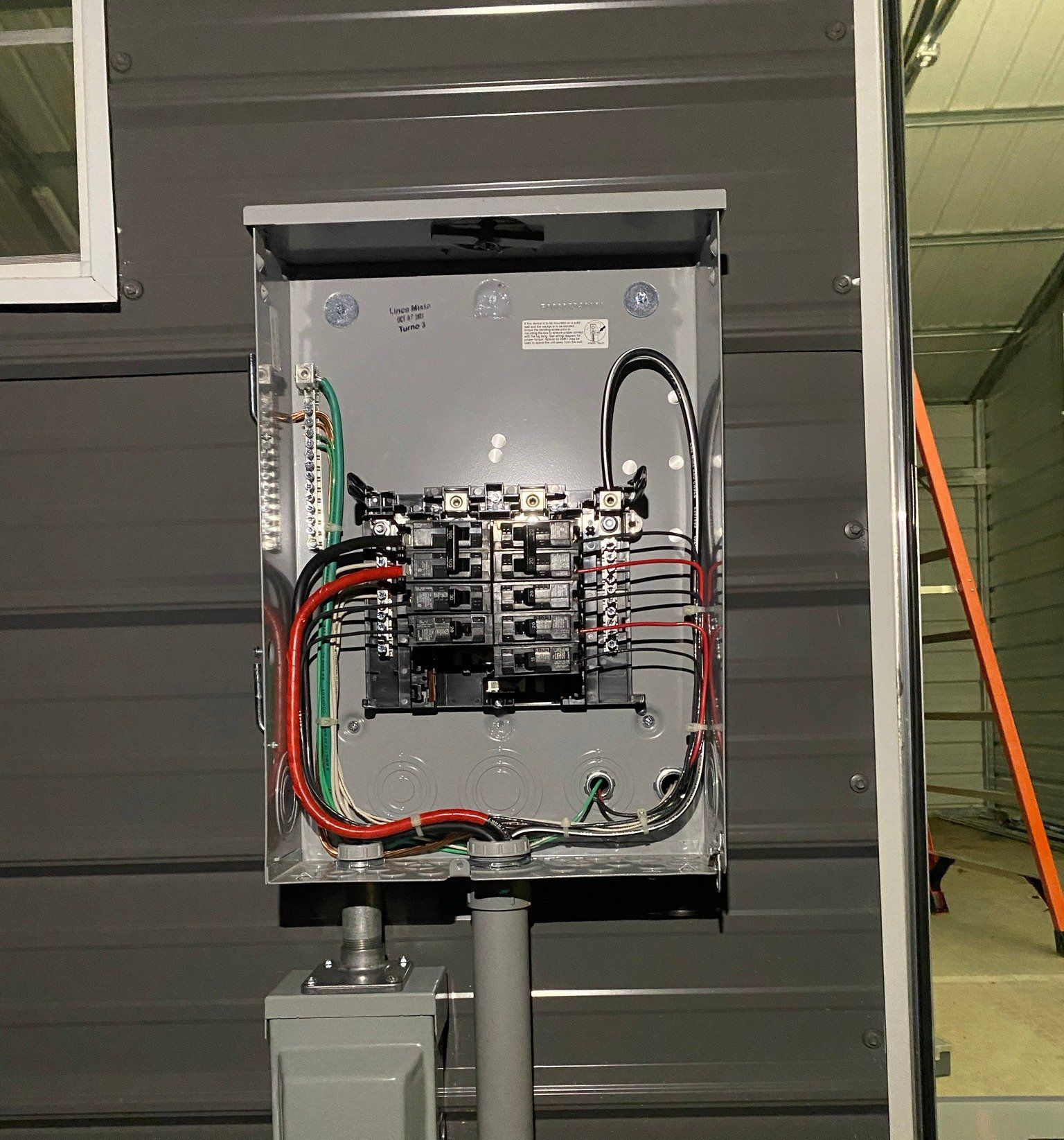 Electrical panel on a gray building exterior, with wiring and breakers visible. Conduit and ground wires are also visible.