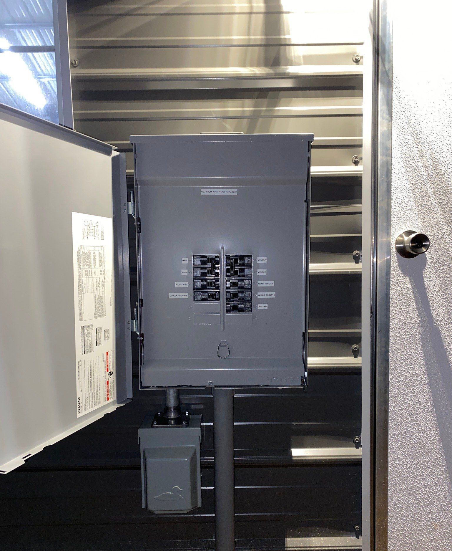 Gray electrical panel with open cover, mounted on a pole, against a corrugated metal wall.