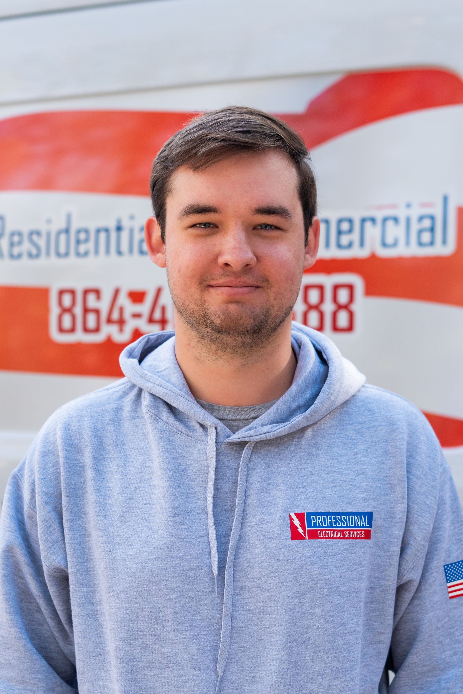 Man in a gray hoodie smiles, standing in front of a service vehicle with company logo and phone number.