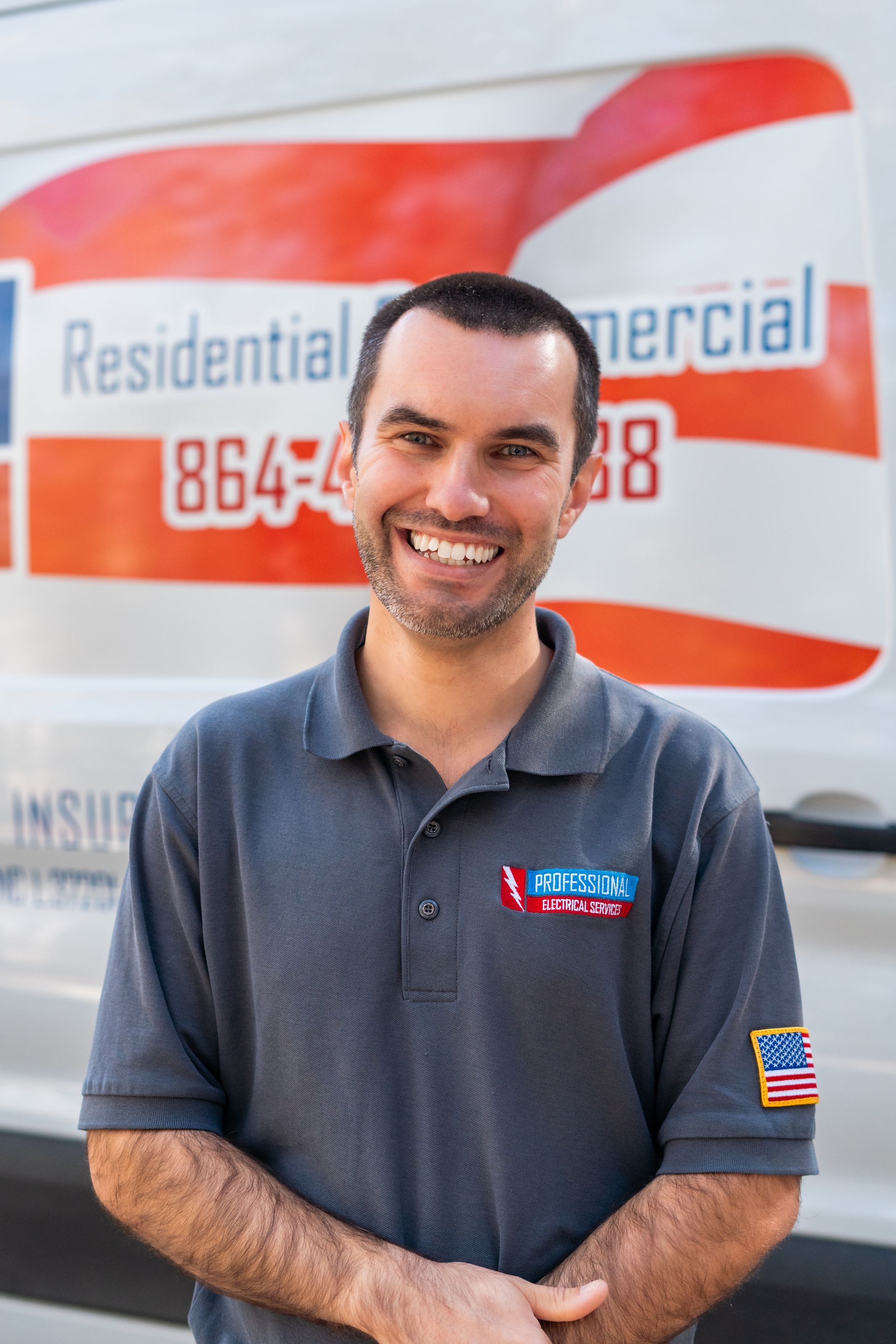 Man smiling, standing in front of a white van with business logo; grey shirt, blue and red accents.