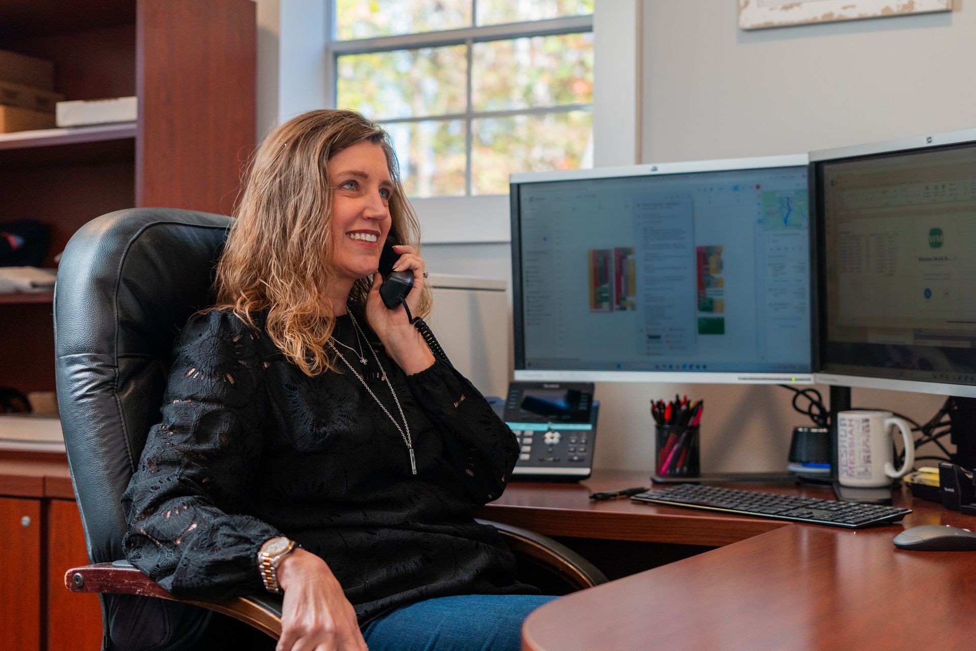 Woman seated at desk, on phone, smiling. Two computer monitors, window in background.