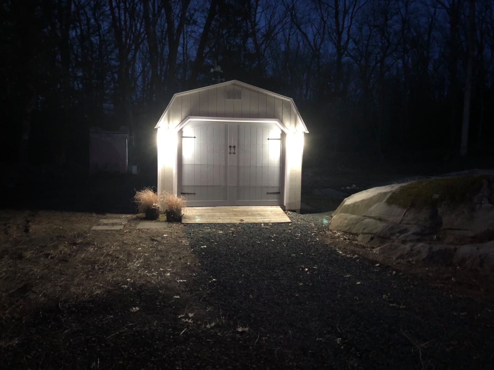 White shed illuminated at night, gravel path leading to it, dark woods in background.