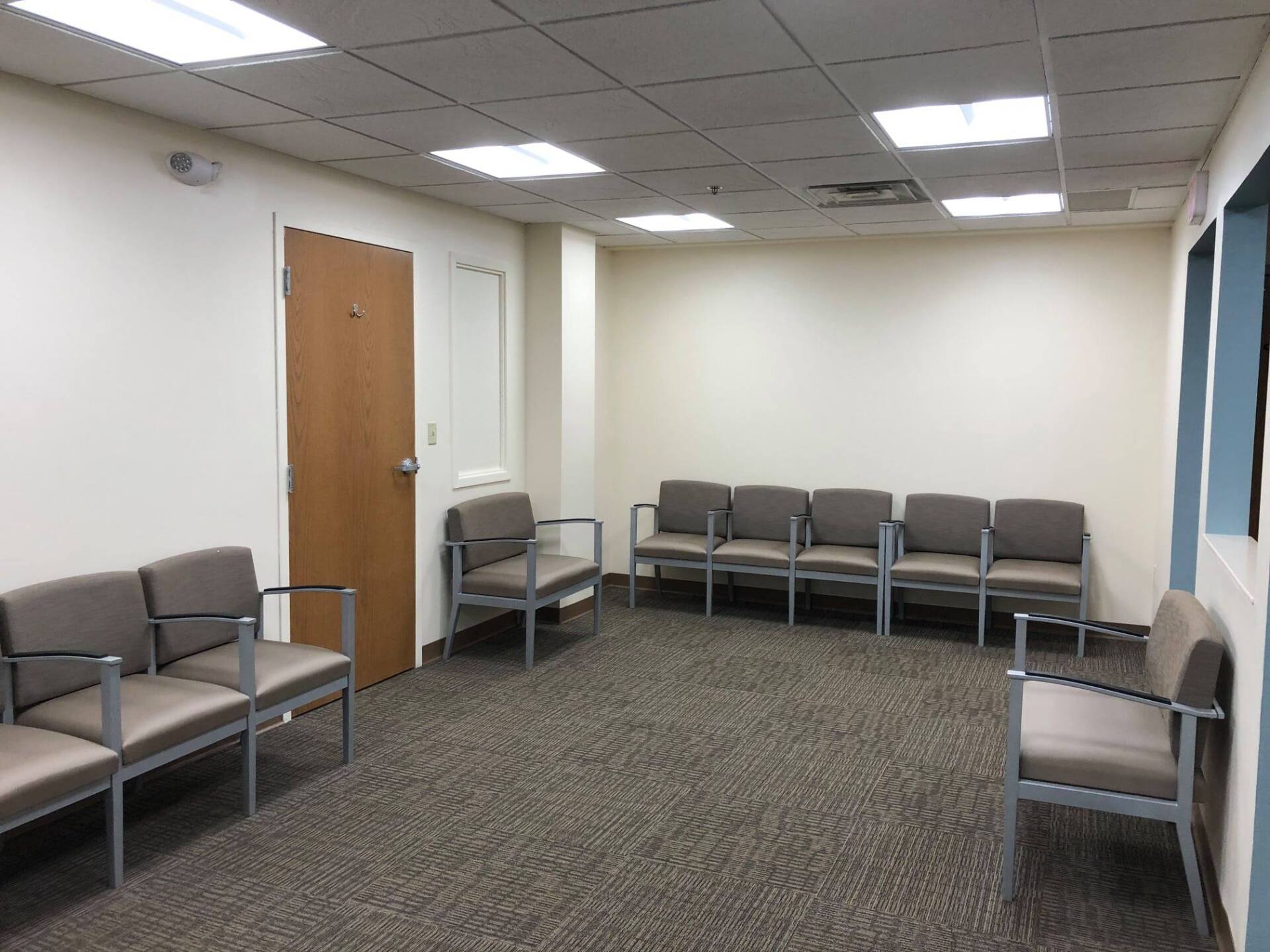 Waiting room with rows of gray chairs, beige walls, brown door, and carpet.