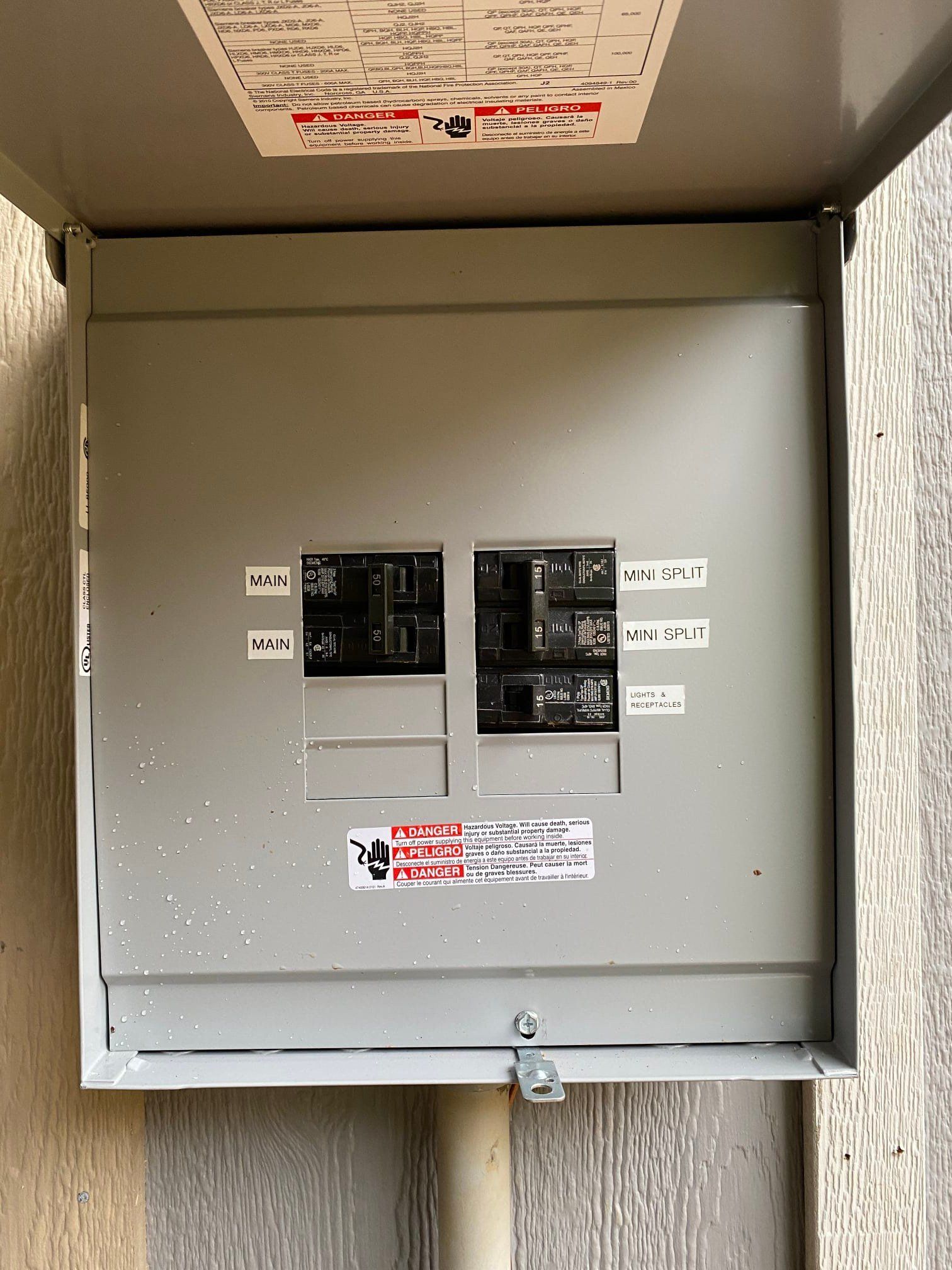 Electrical panel with labels, breakers, and a closed metal door, mounted on a wall.