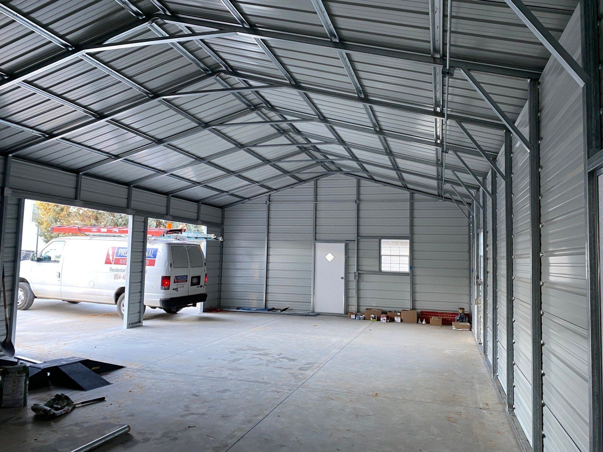 Inside of a metal-framed garage with a vehicle visible outside the open bay. White walls, concrete floor, and a door are also present.