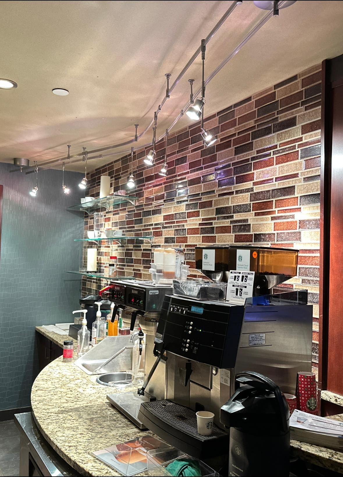 Coffee shop counter with espresso machines, a brick-patterned wall, and track lighting.