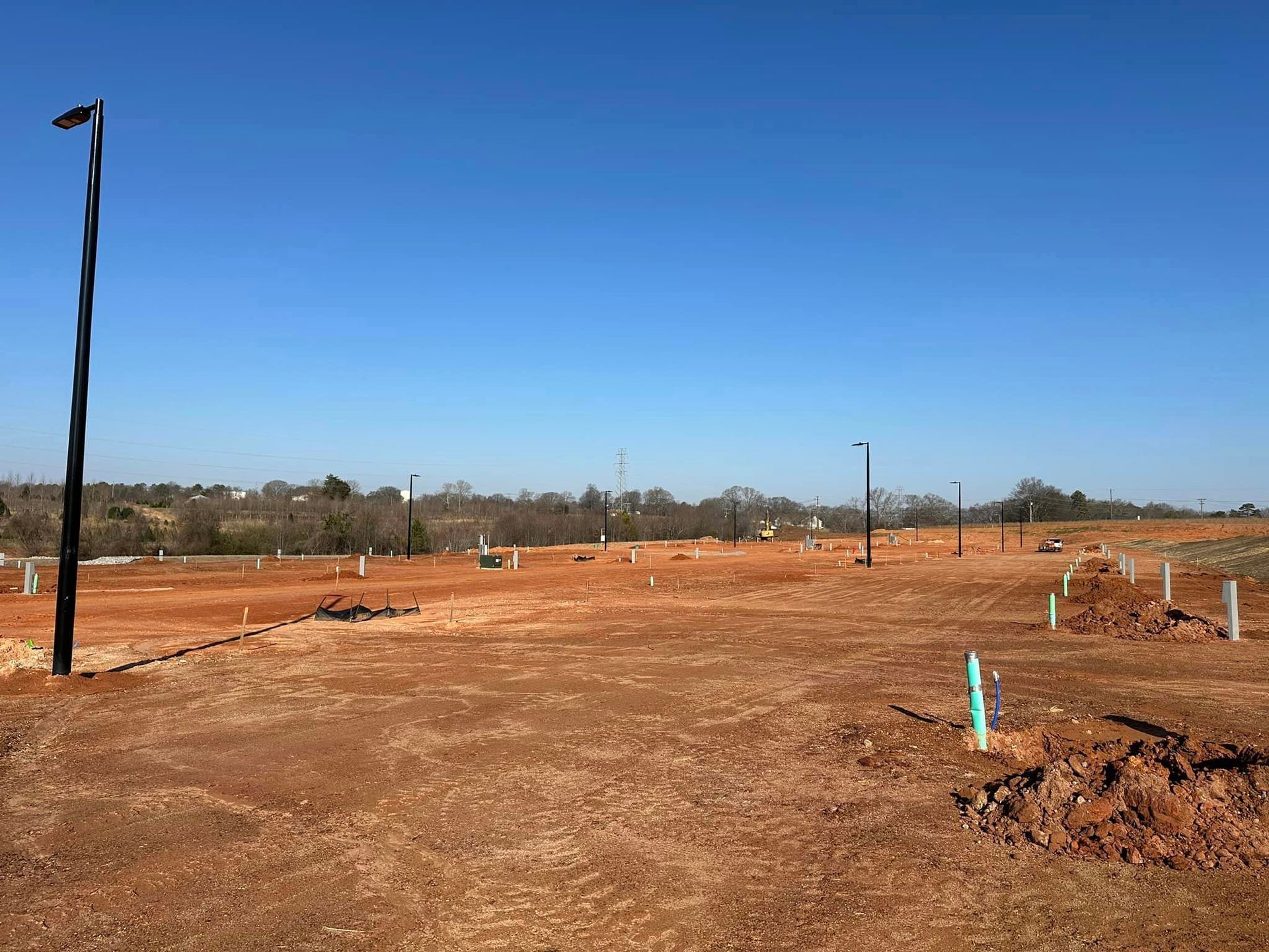 Cleared lot with red soil, utility pipes, and streetlights under a blue sky, distant trees.