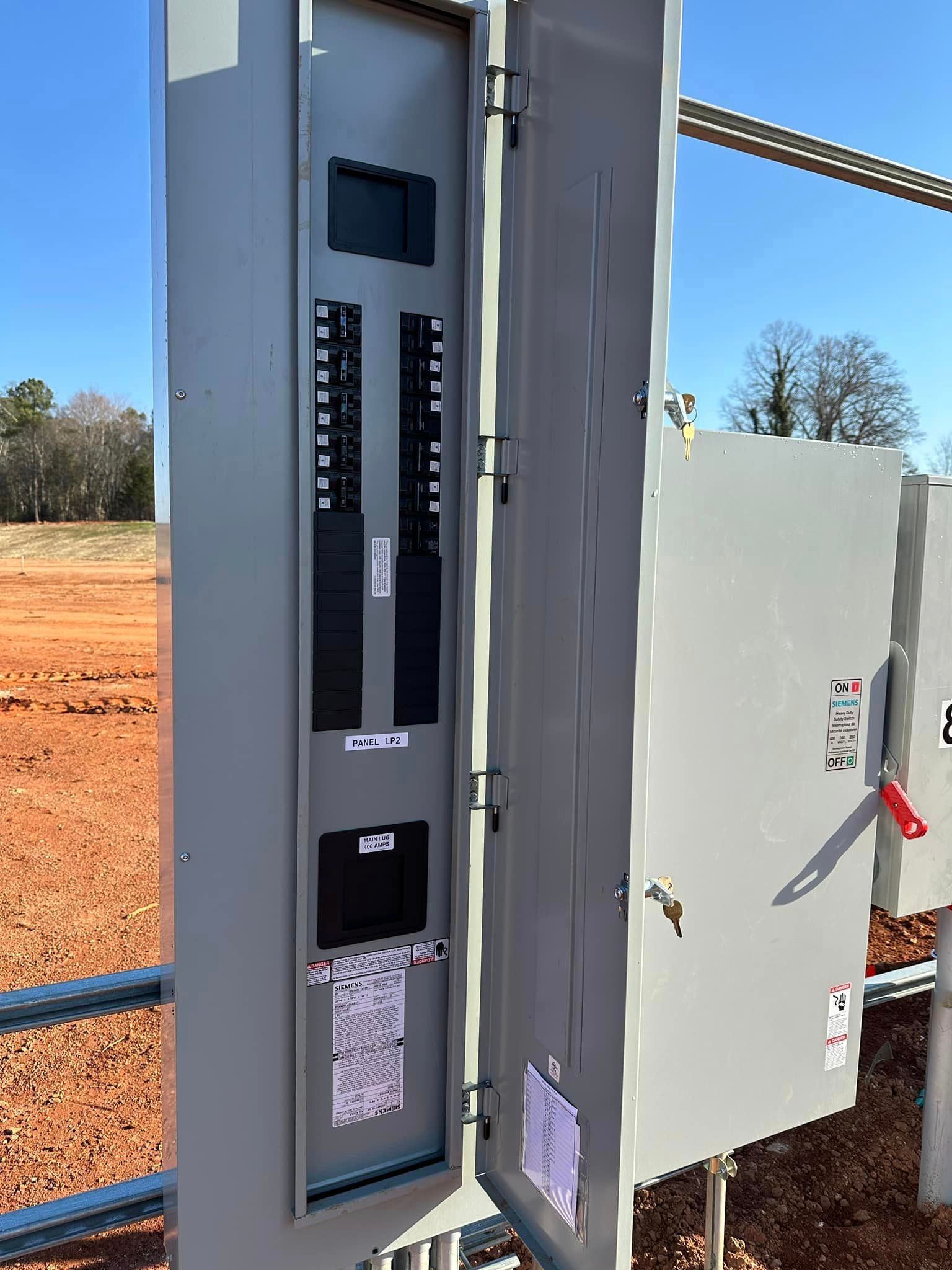 Gray electrical panel box, open, revealing circuit breakers and a label, outdoors in a field.