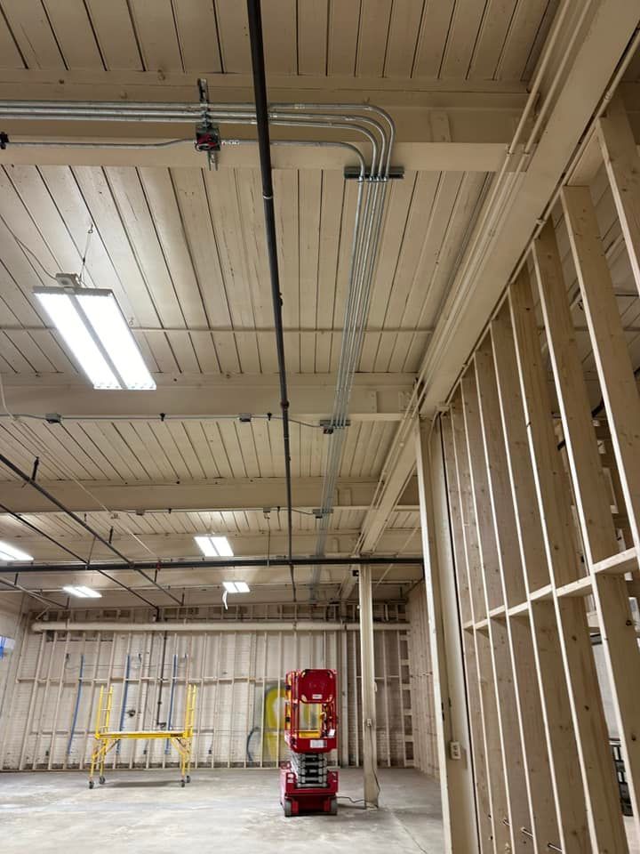 Interior view of a construction site with electrical conduit running across the ceiling. Wooden framing and fluorescent lights are also visible.