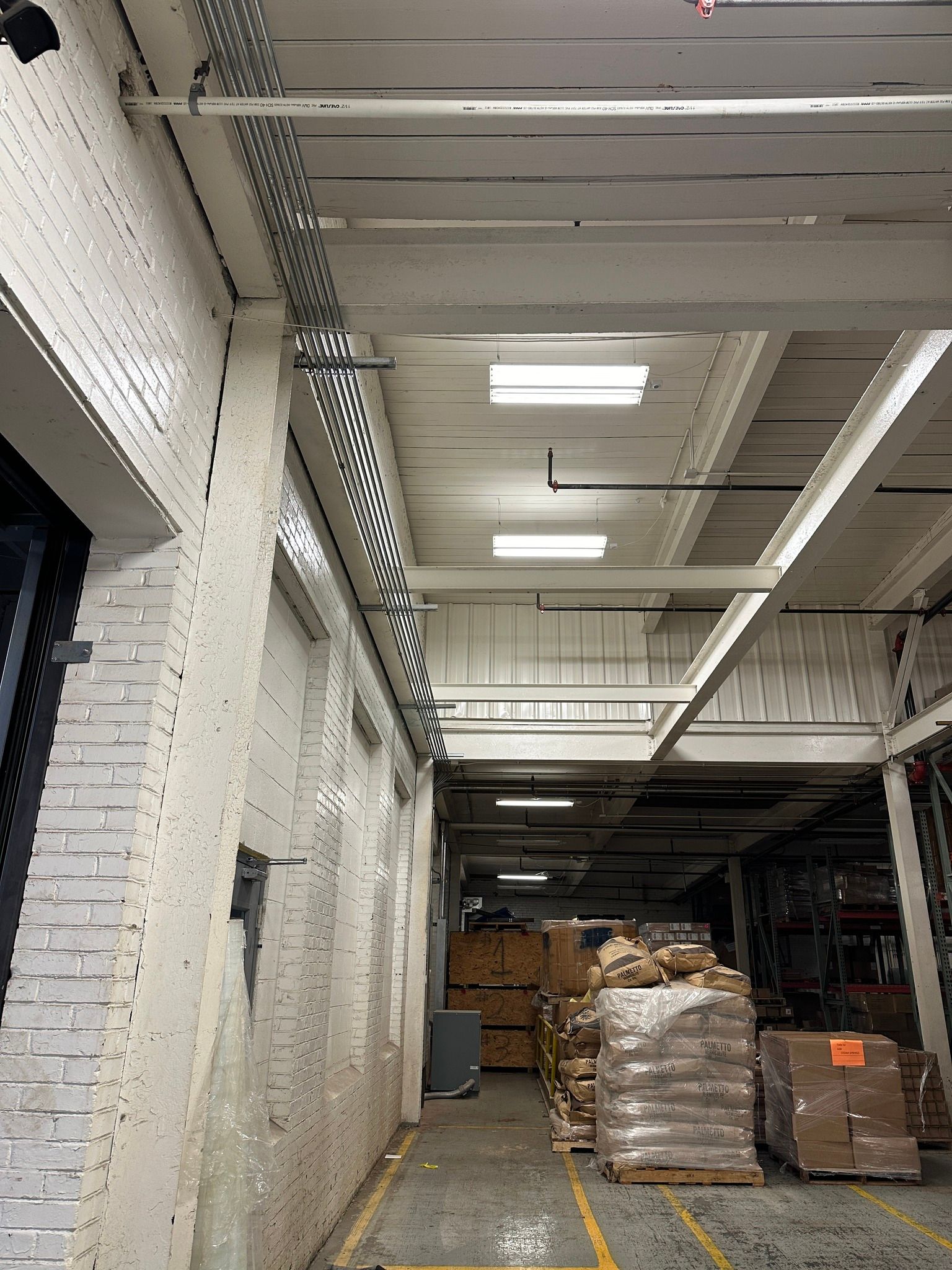 Warehouse interior: White brick wall, metal cable trays, overhead lighting, pallets of goods.