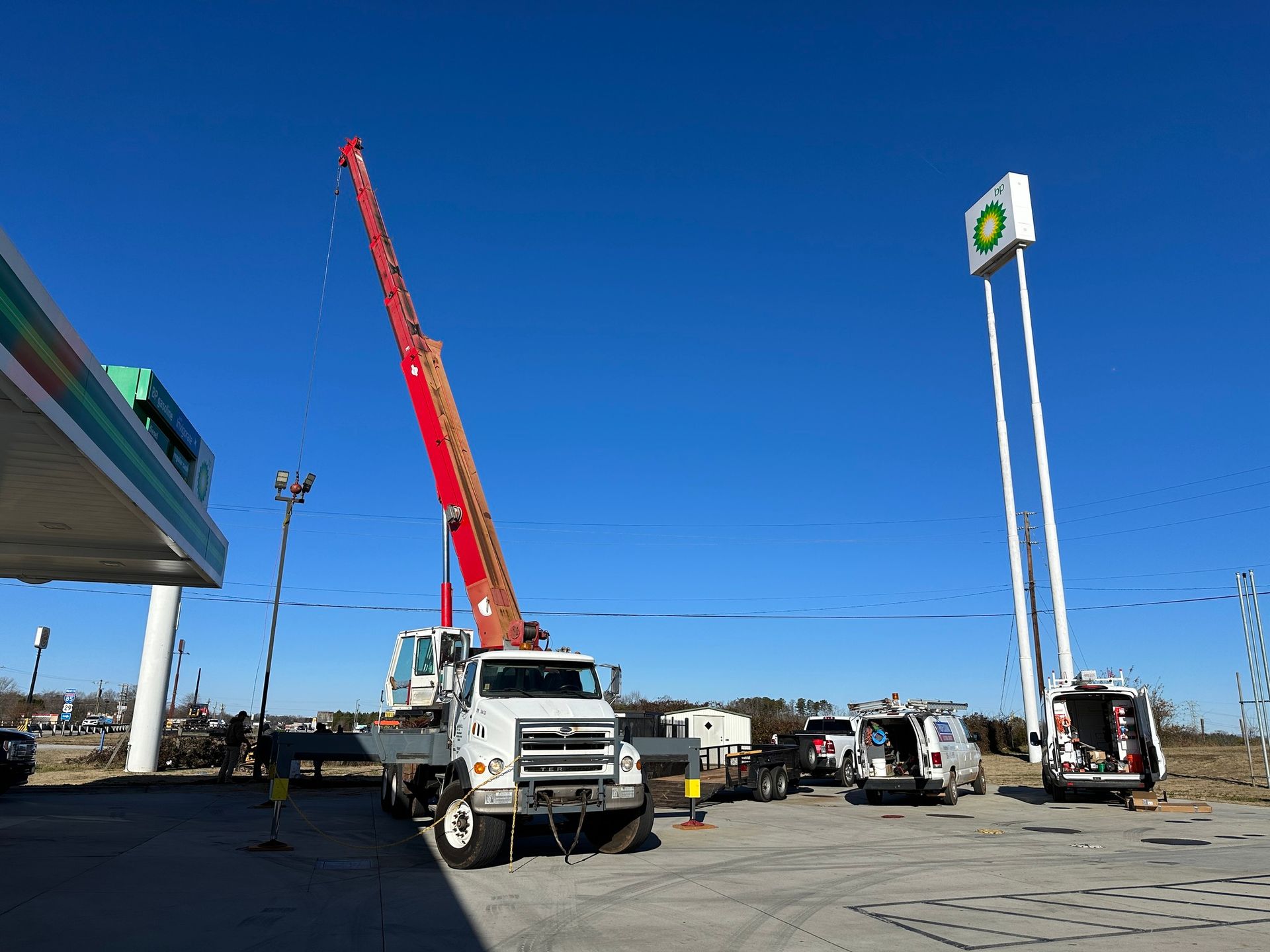 Crane truck near a BP gas station, extending its boom towards a tall sign post. Bright blue sky.