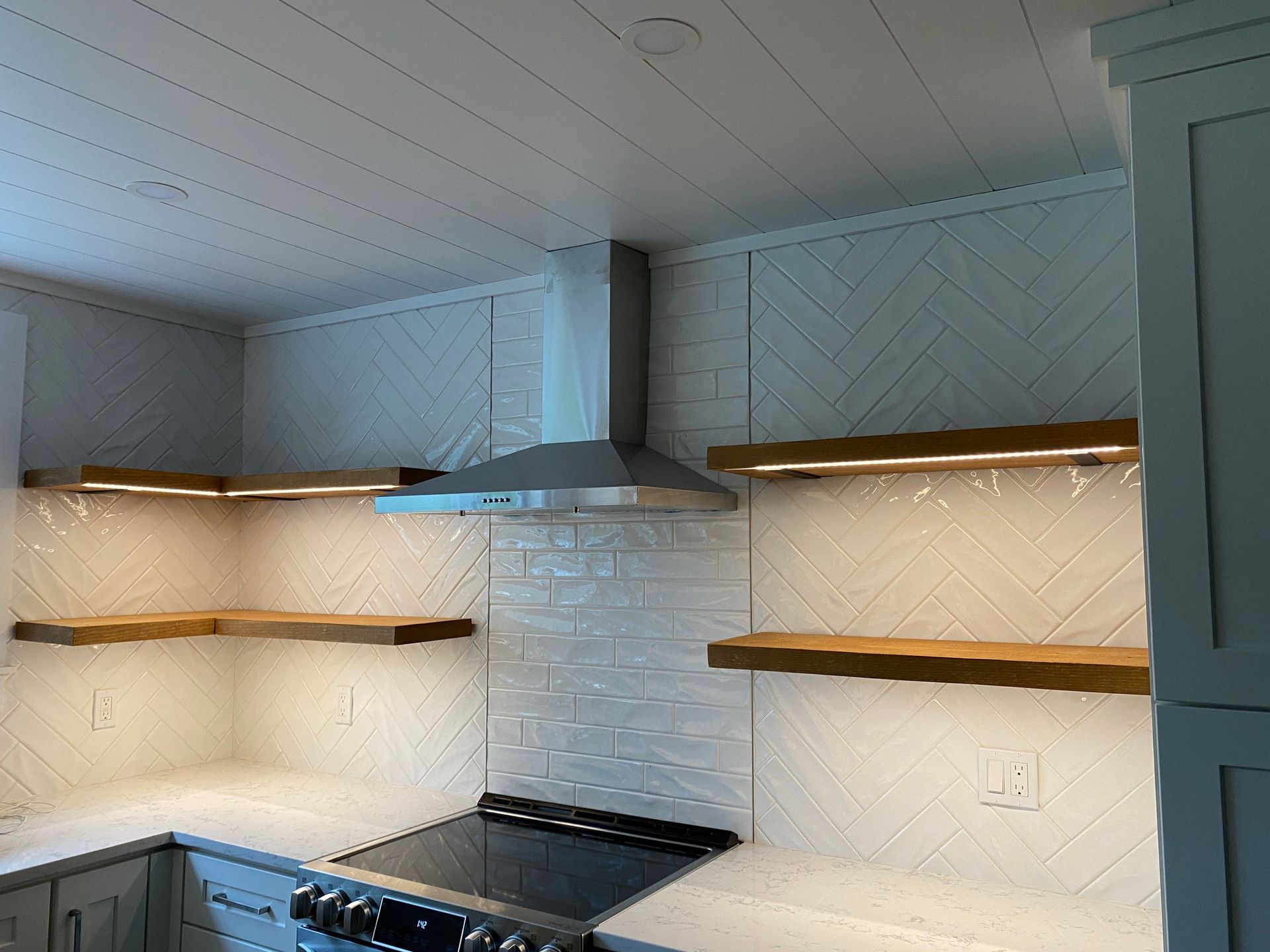Kitchen with wooden shelves, white backsplash, range hood, and stove. Shelves have under-cabinet lighting.