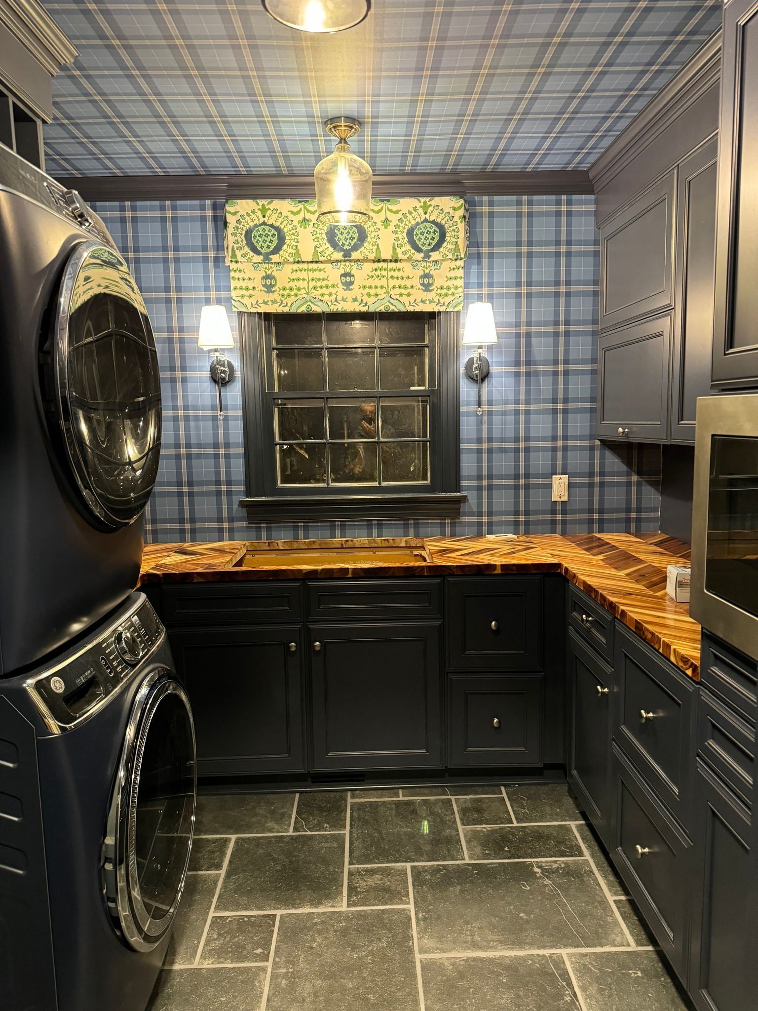 Laundry room with dark blue cabinets, plaid wallpaper, stacked washer/dryer, stone floor, and a wooden countertop.