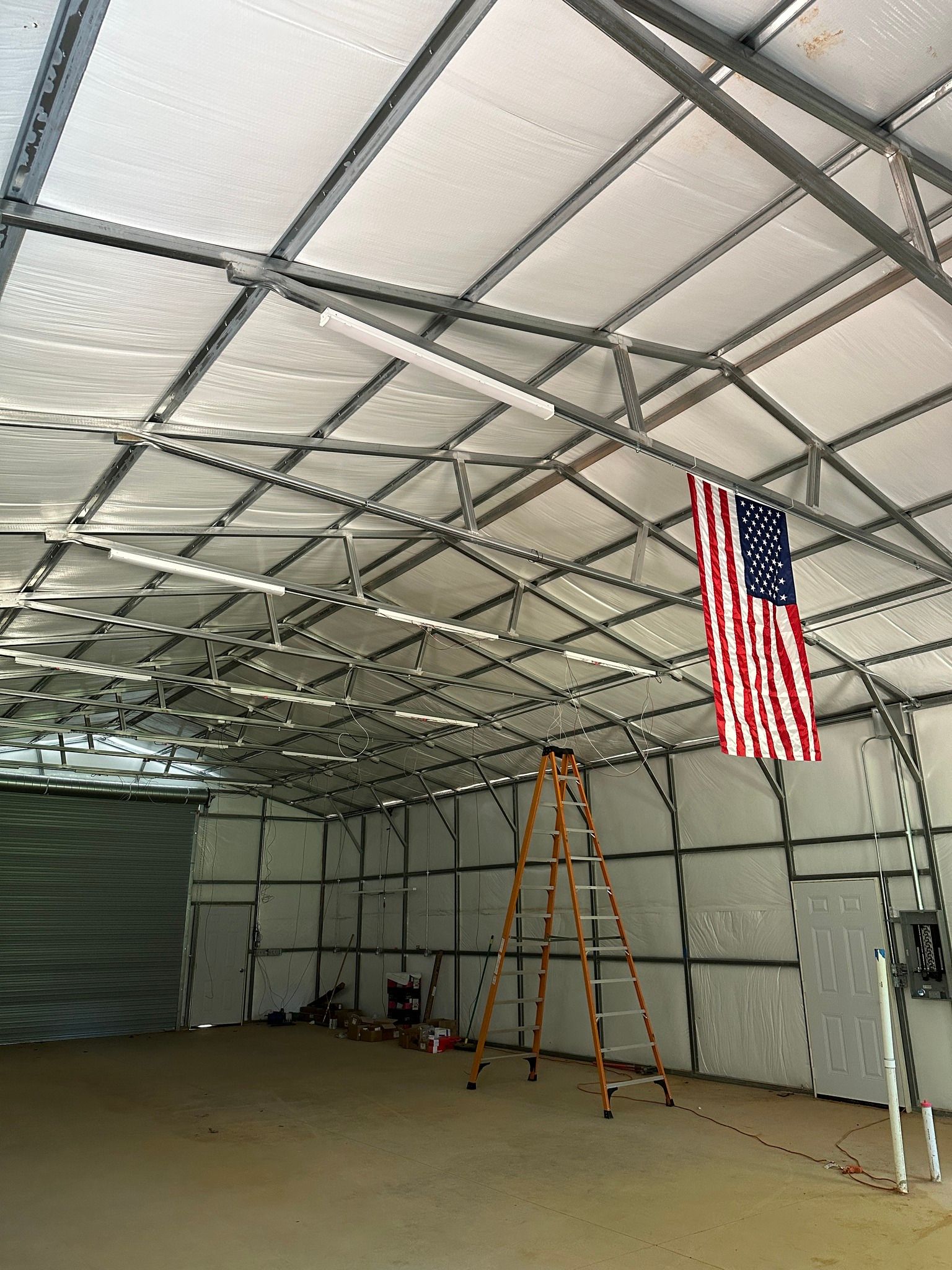Inside a metal-framed building: an American flag hangs from the ceiling; a ladder stands below.