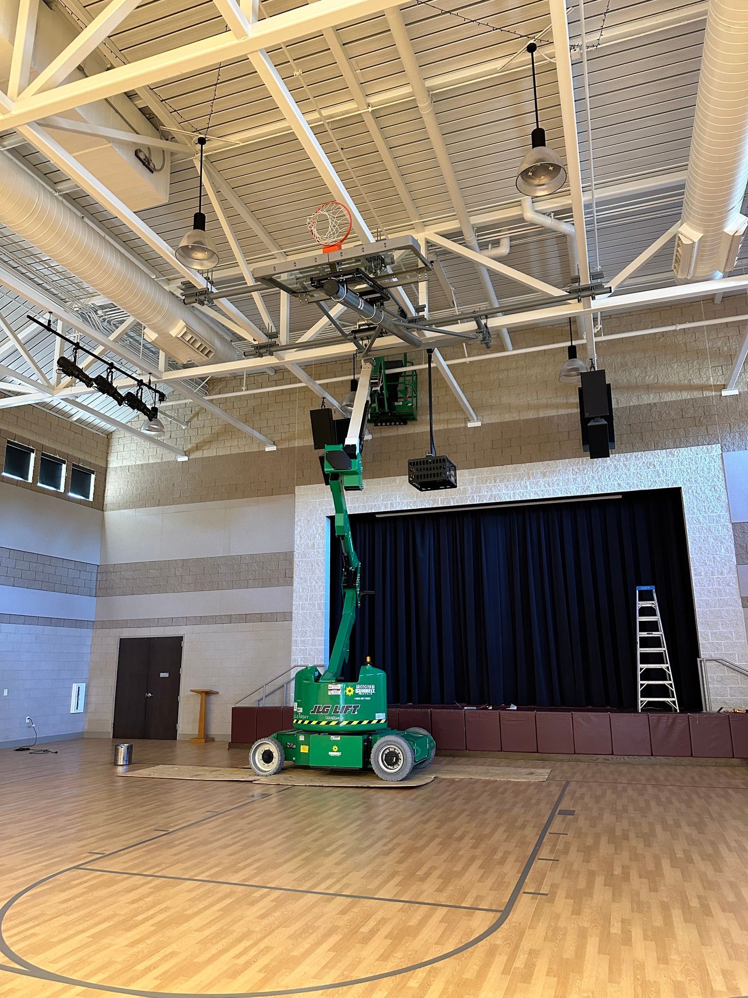 A green lift in a gymnasium adjusts overhead lighting and speakers. Stage with black curtain and basketball court.