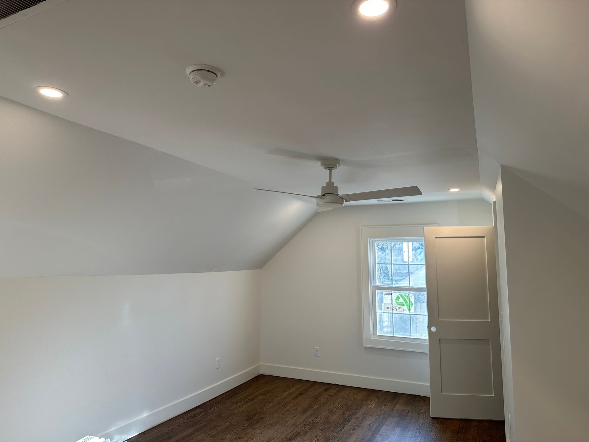 Empty room with sloped ceiling, ceiling fan, window, white walls, and hardwood floor.