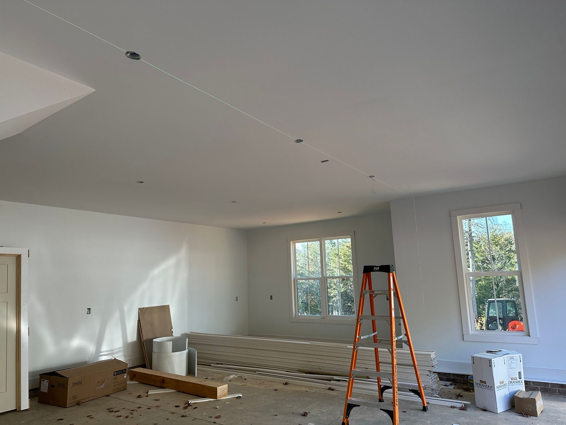 Interior of a room under construction with white walls and ceiling; drywall and a ladder are present.