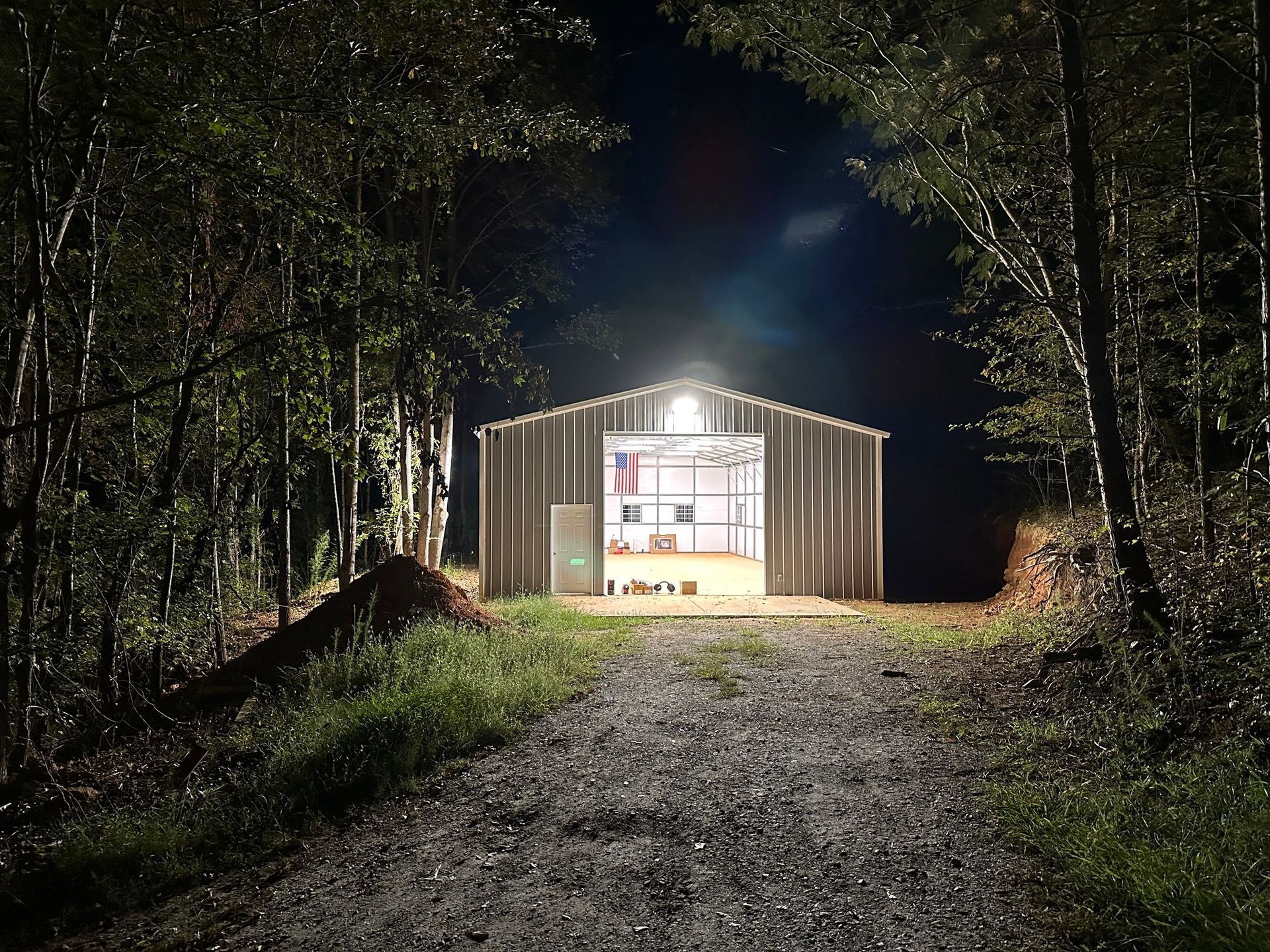 A metal shed glows at night, illuminated by a bright overhead light, at the end of a gravel path in a forest.