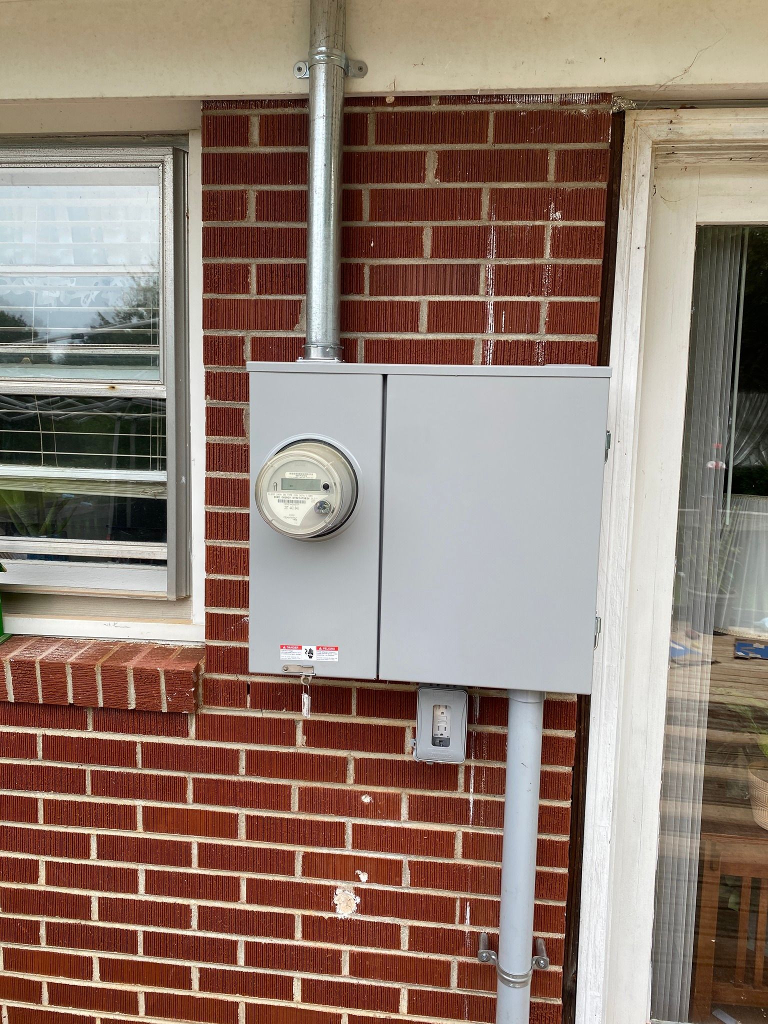 Electric meter and panel box mounted on a red brick wall next to a window and door.
