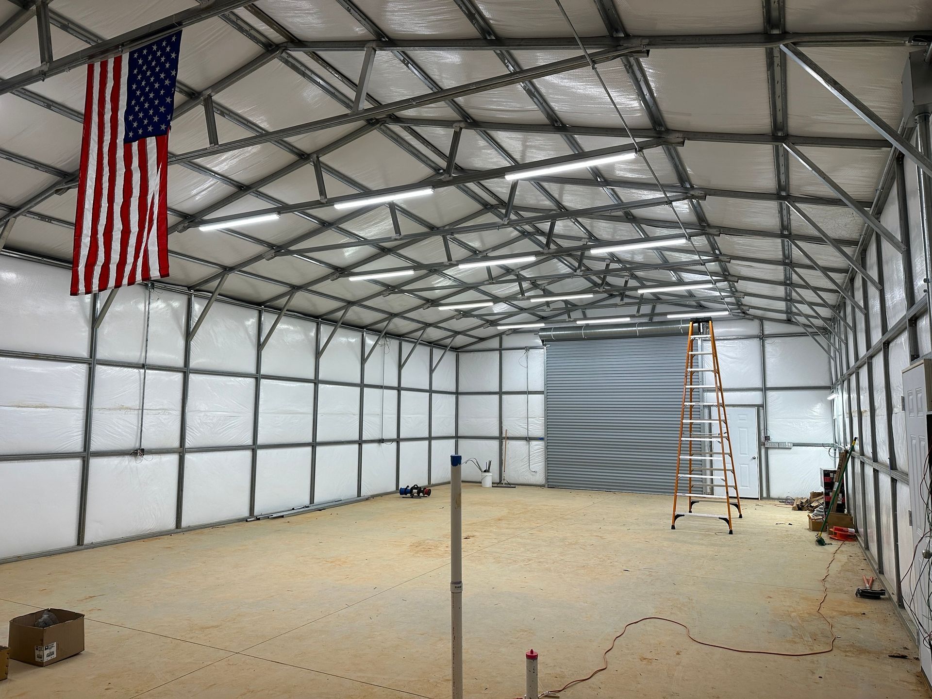 Large, empty metal-framed building interior with American flag, a ladder, and overhead lights.