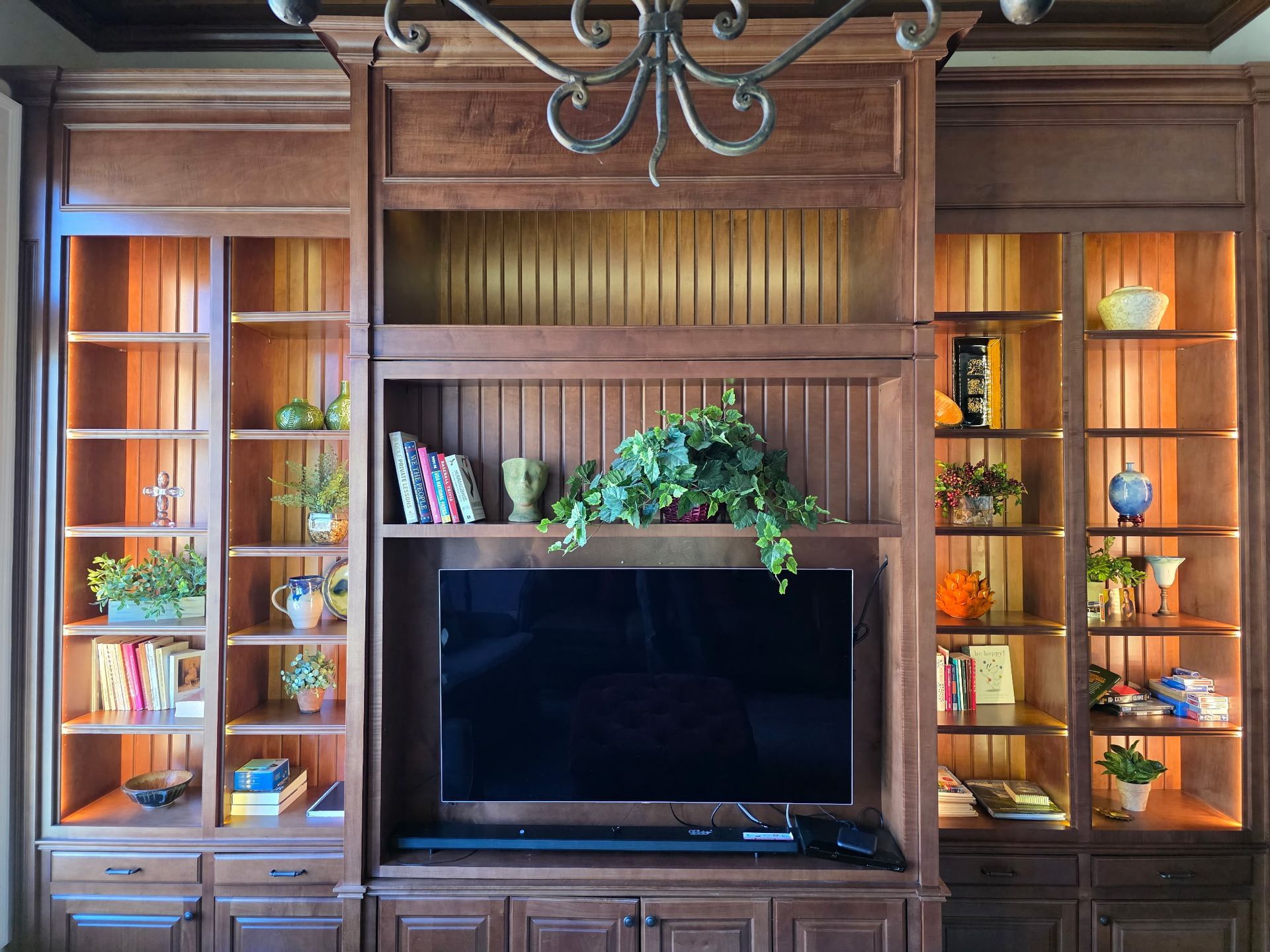 Built-in wooden entertainment center with shelves, TV, and decorative items. Warm lighting, brown wood, and a chandelier.