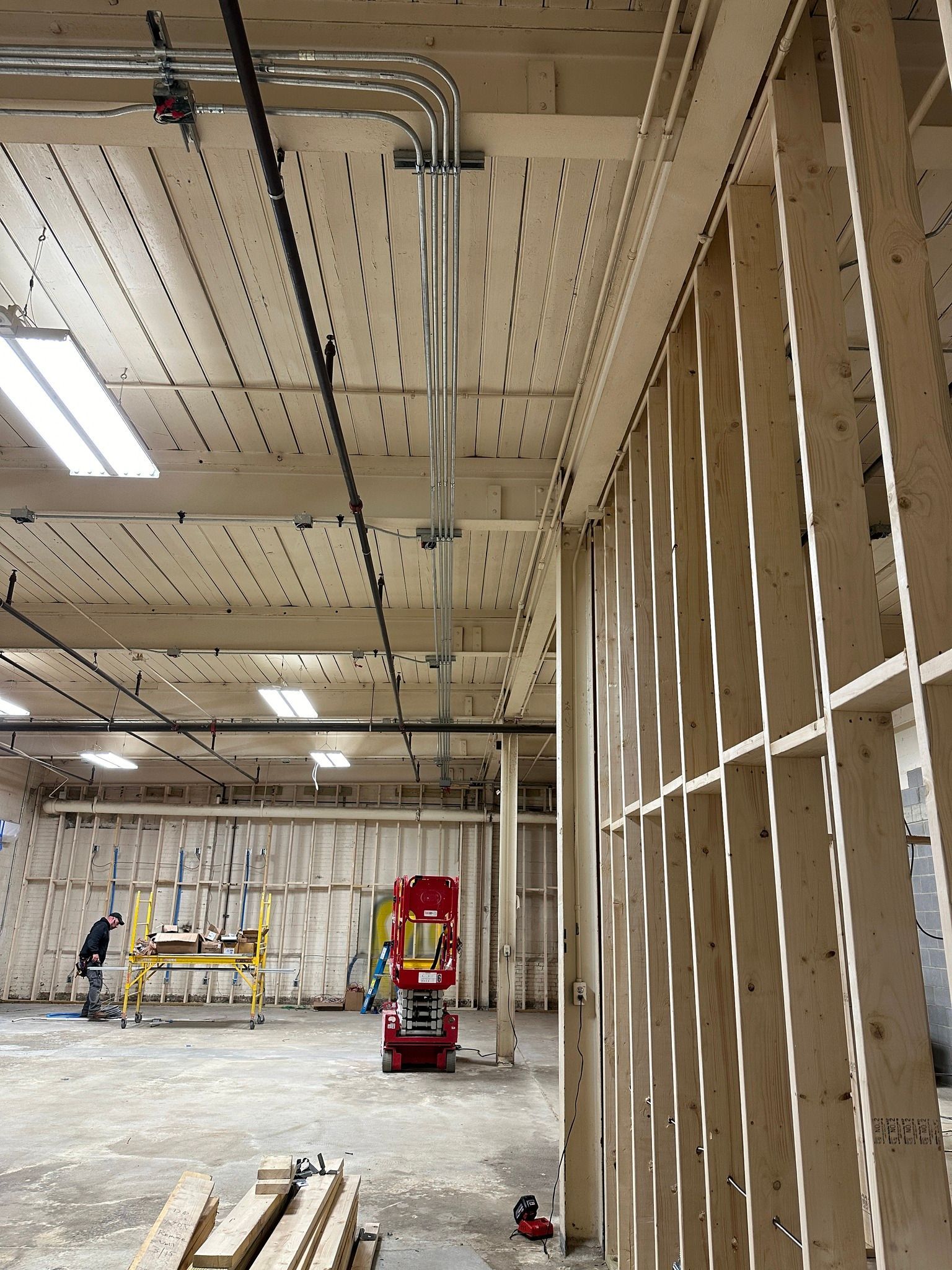 Interior construction site: wooden framing, exposed ceiling with electrical conduit, concrete floor, workers, and equipment.
