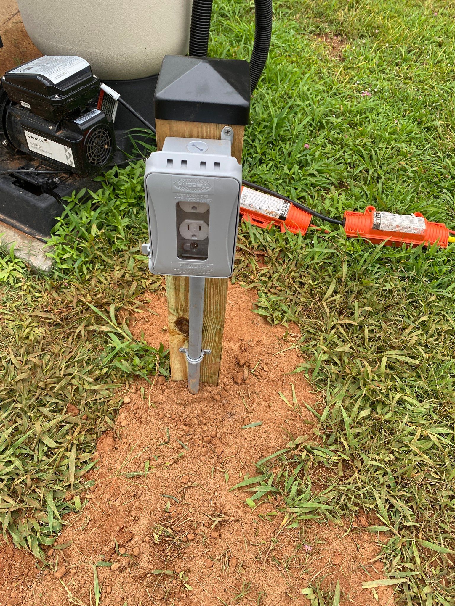Gray electrical outlet box on a wooden post in a grassy yard, with connected orange extension cords.