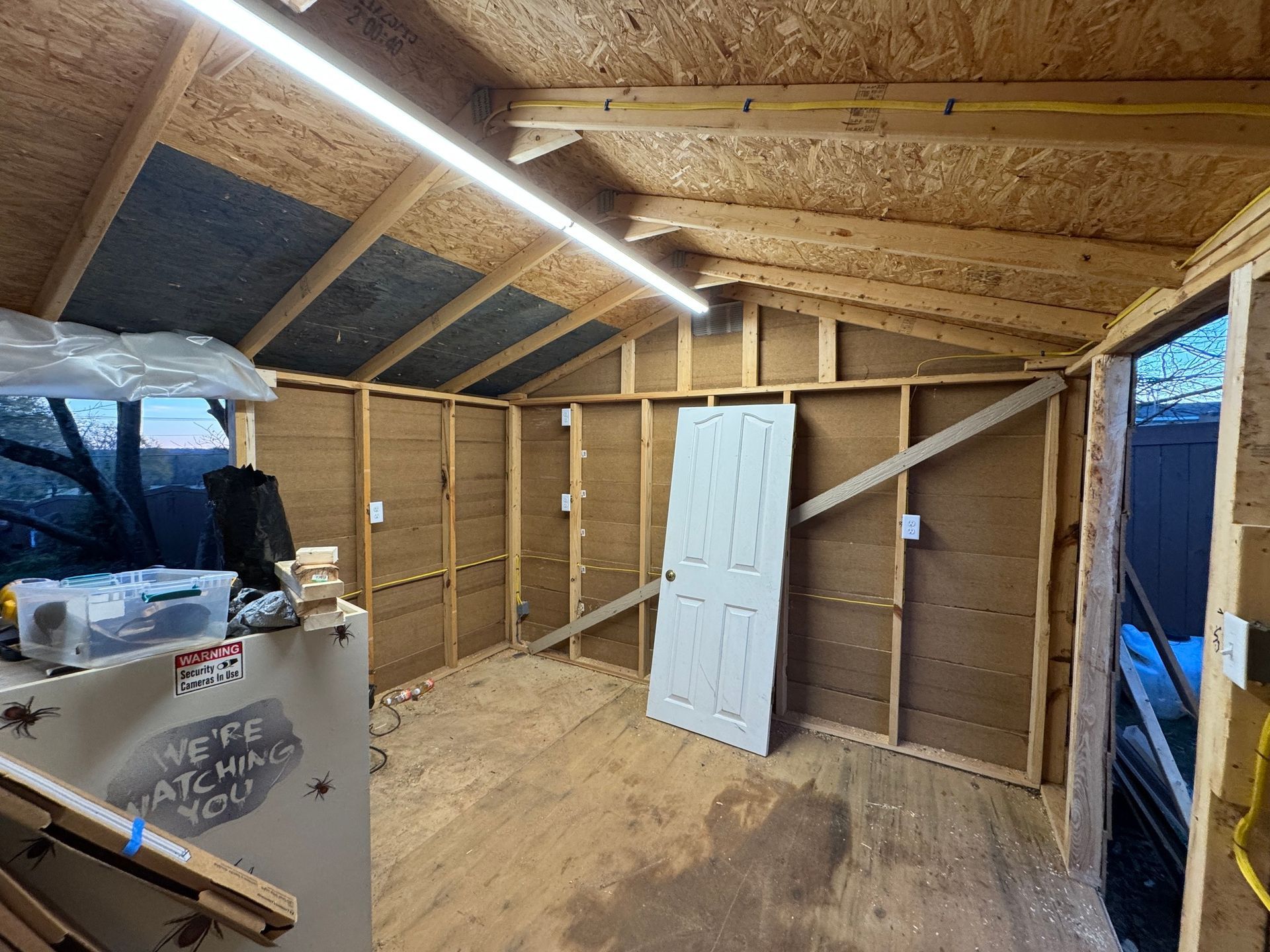 Interior of a wood-framed shed under construction with door, exposed studs, and light fixtures.