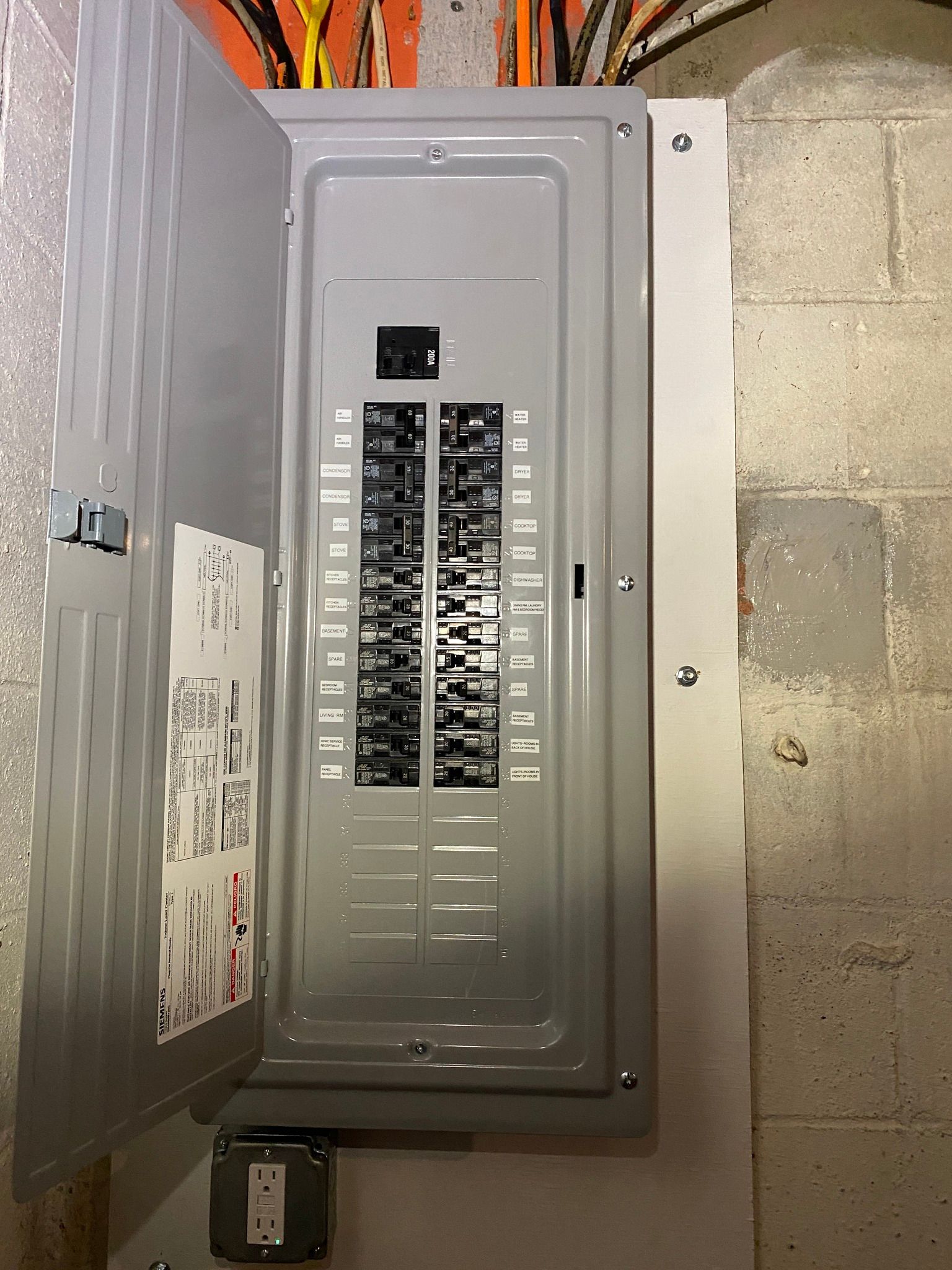 Opened gray electrical panel box in a basement setting, revealing circuit breakers.